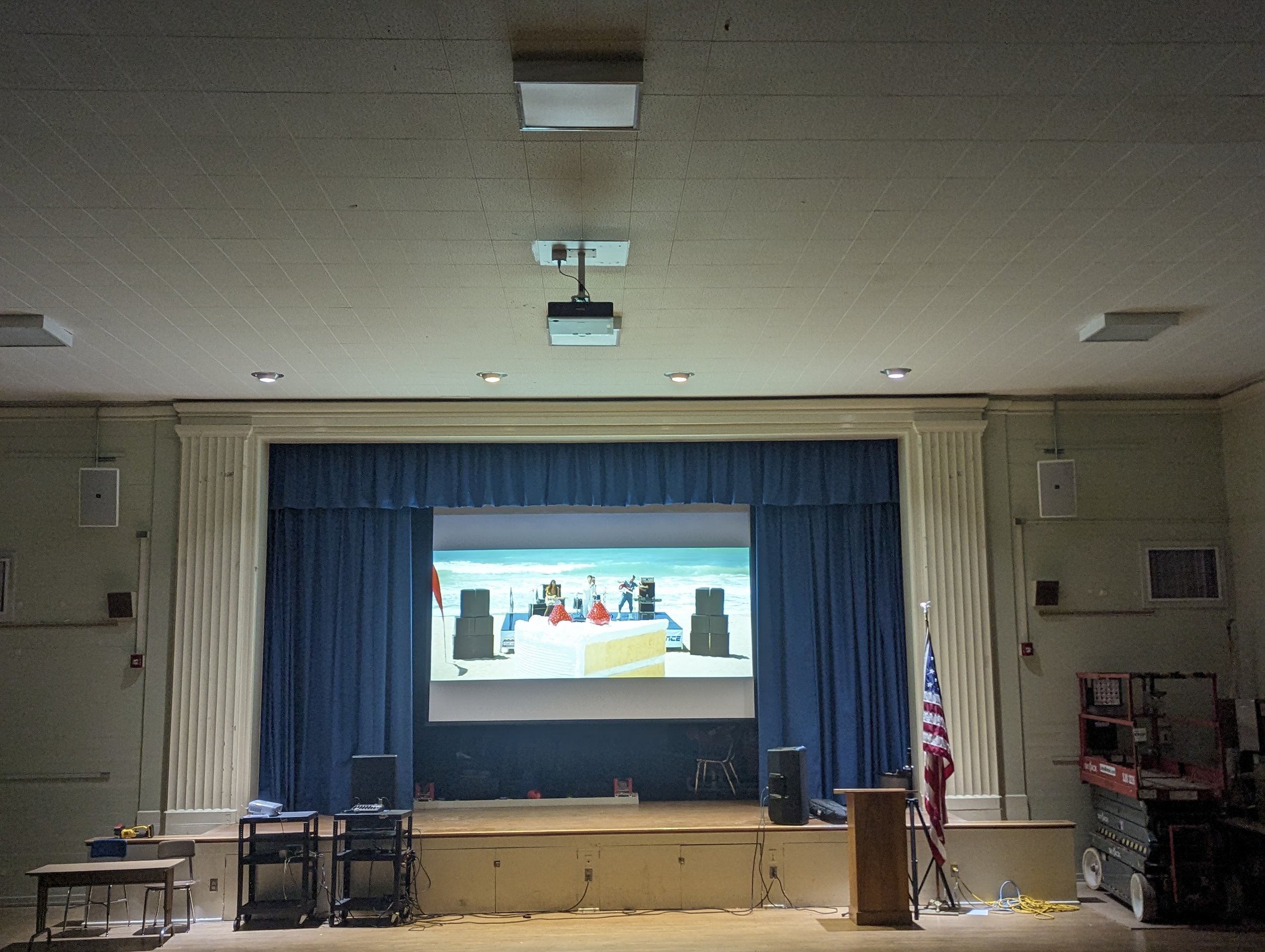 An empty auditorium stage with a large screen showing a beach scene with musical instruments and speakers, framed by blue curtains, and an American flag standing to the right of the stage.