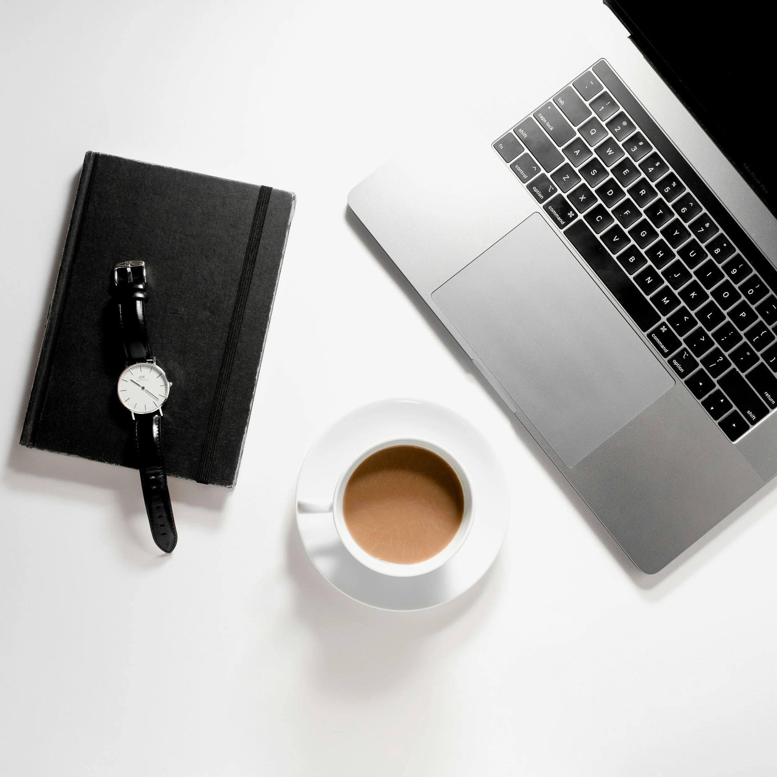 Overhead view of a workspace with a silver laptop, a black notebook with a black watch on top, a white cup of coffee, and a black thick pen on the notebook, all on a white surface.
