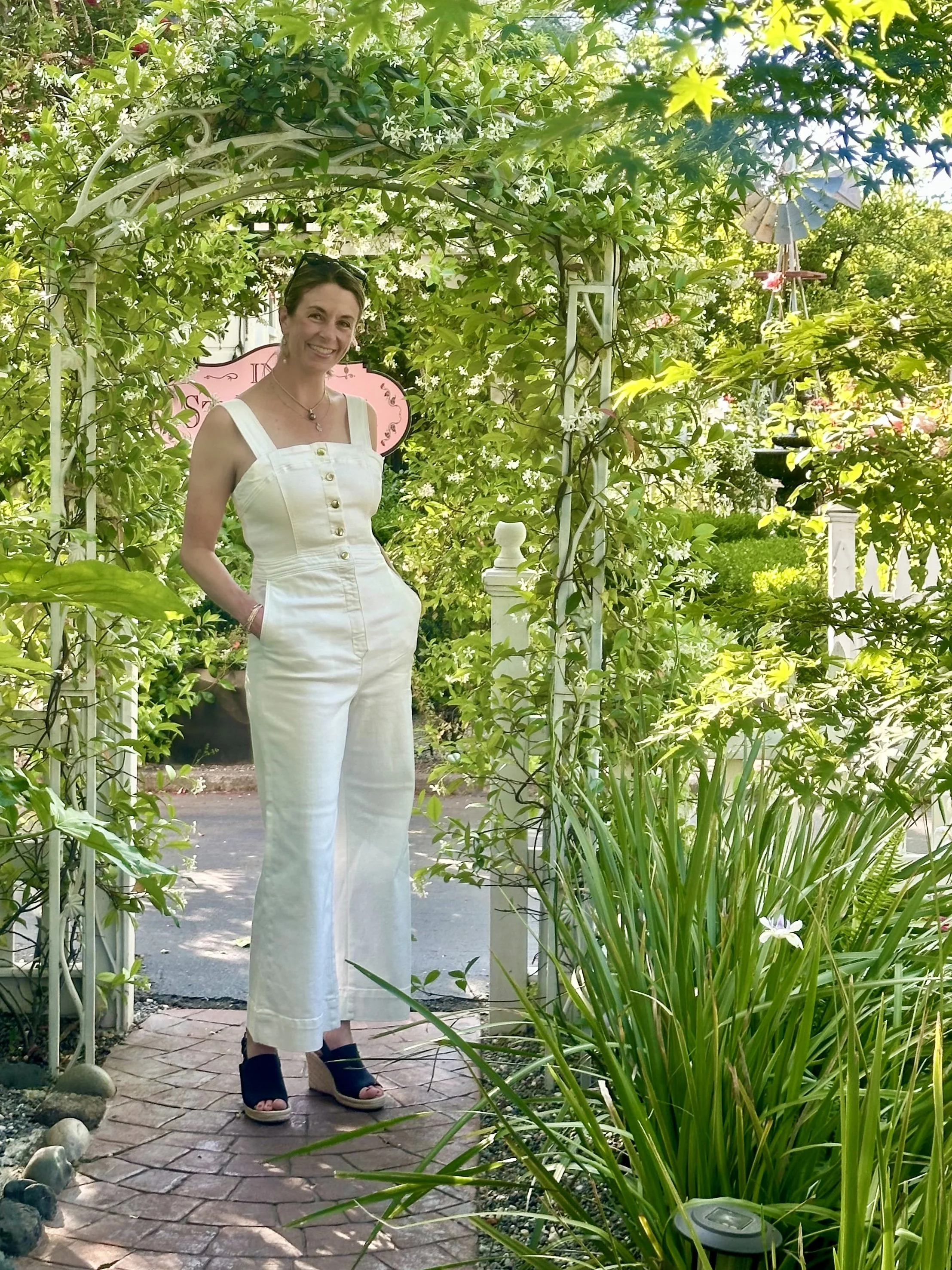 A woman in a white sleeveless jumpsuit standing on a brick pathway in a lush garden archway surrounded by green plants and flowers, smiling at the camera.