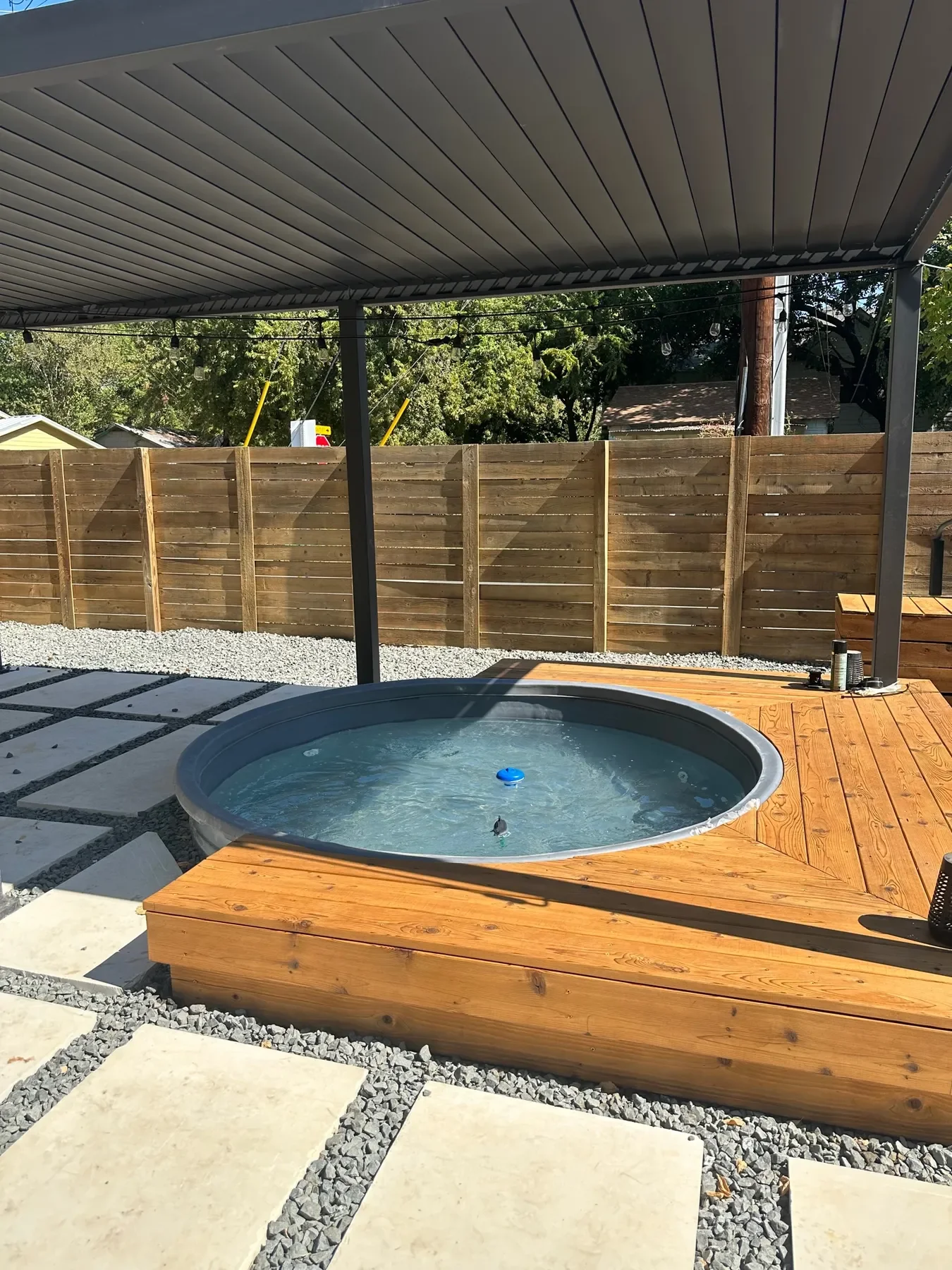 An outdoor backyard hot tub on a wooden deck beneath a metal canopy, with a wooden fence and trees in the background.