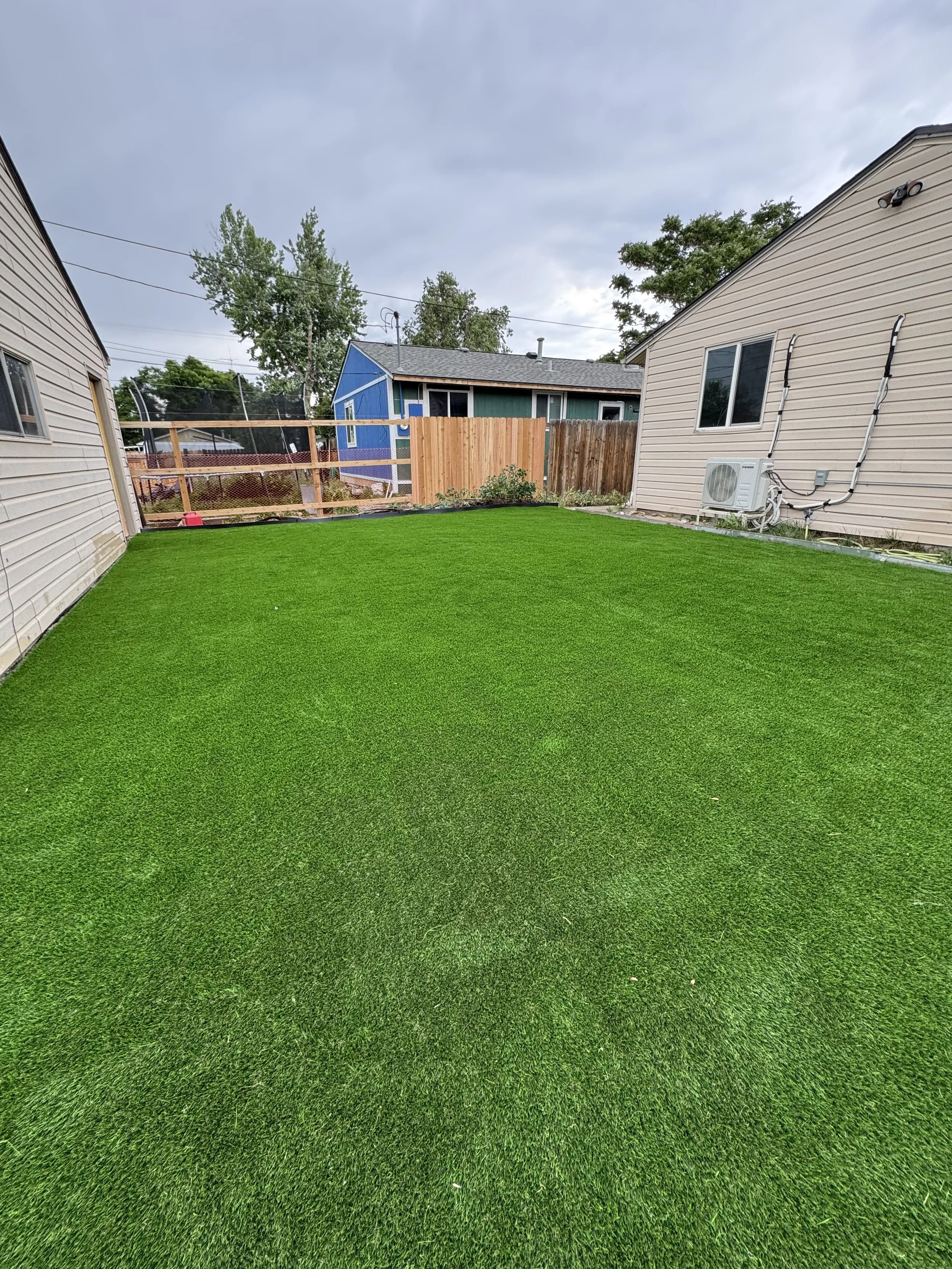 View of a backyard with bright green artificial grass, beige and blue houses, and a cloudy sky.