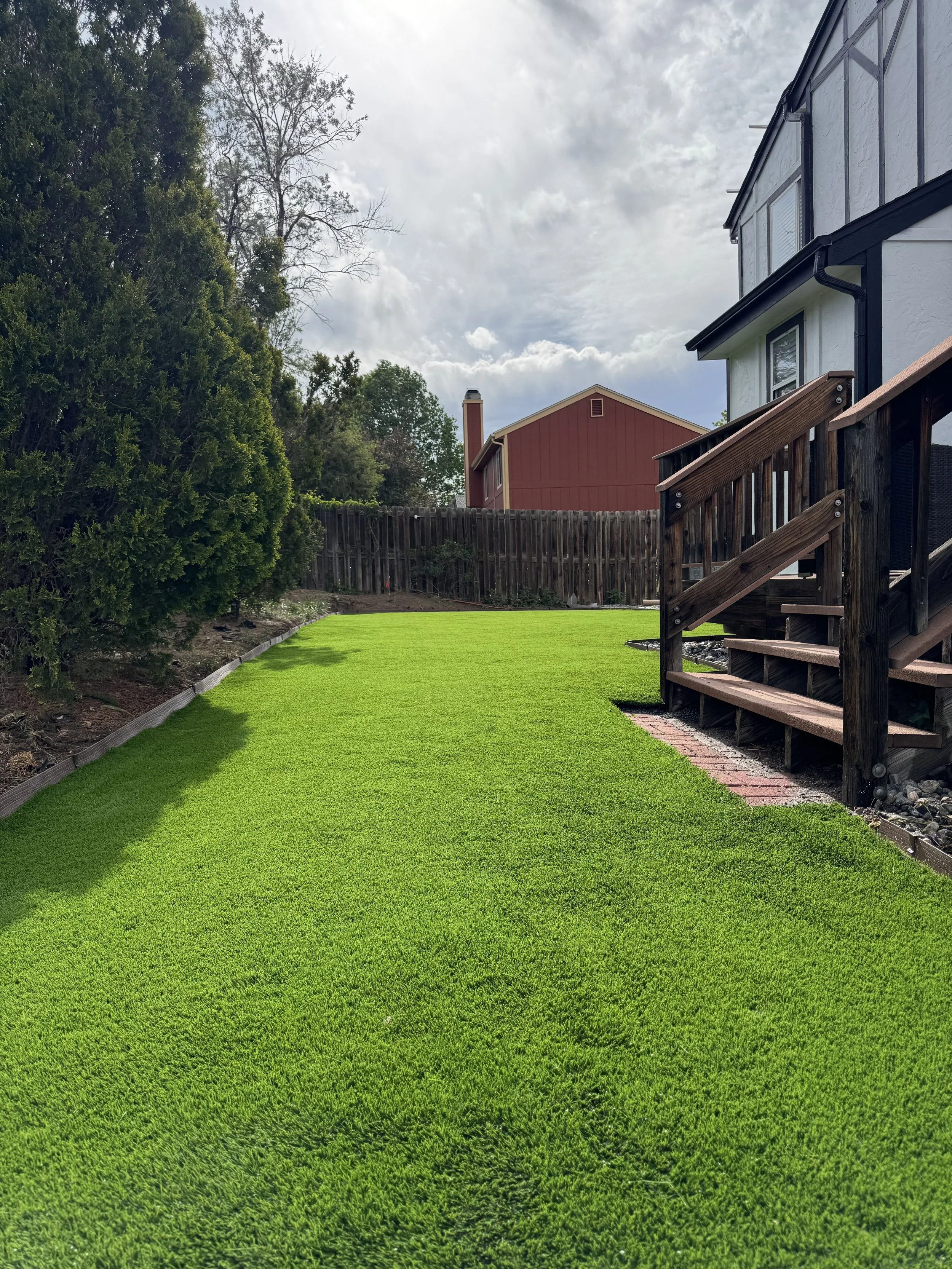 Well-manicured green grass lawn with a small brick pathway and wooden steps leading to a raised porch, surrounded by trees and a fence, under a partly cloudy sky.