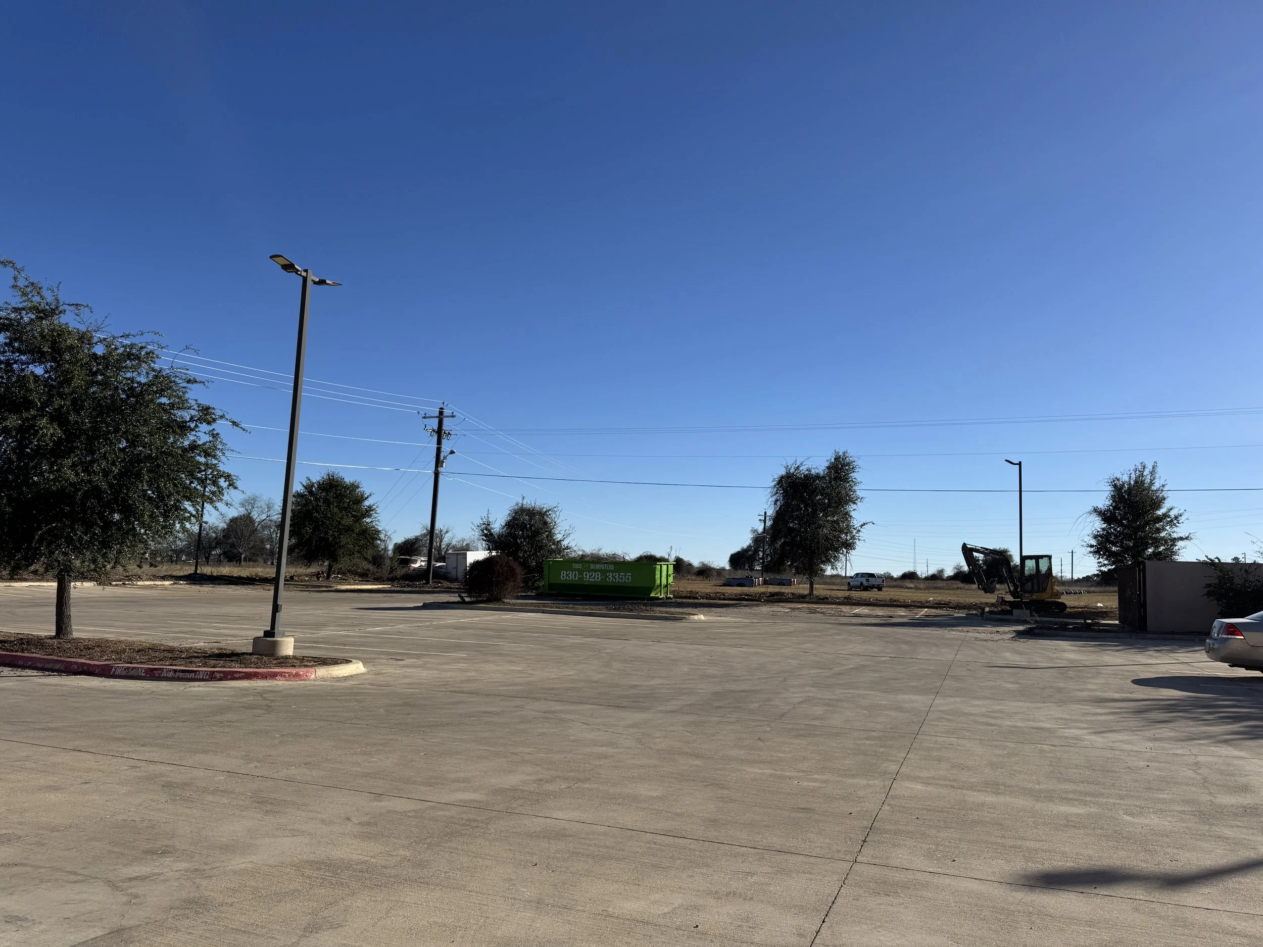Empty parking lot with a few trees, a green dumpster, construction equipment, and utility poles under a clear blue sky.