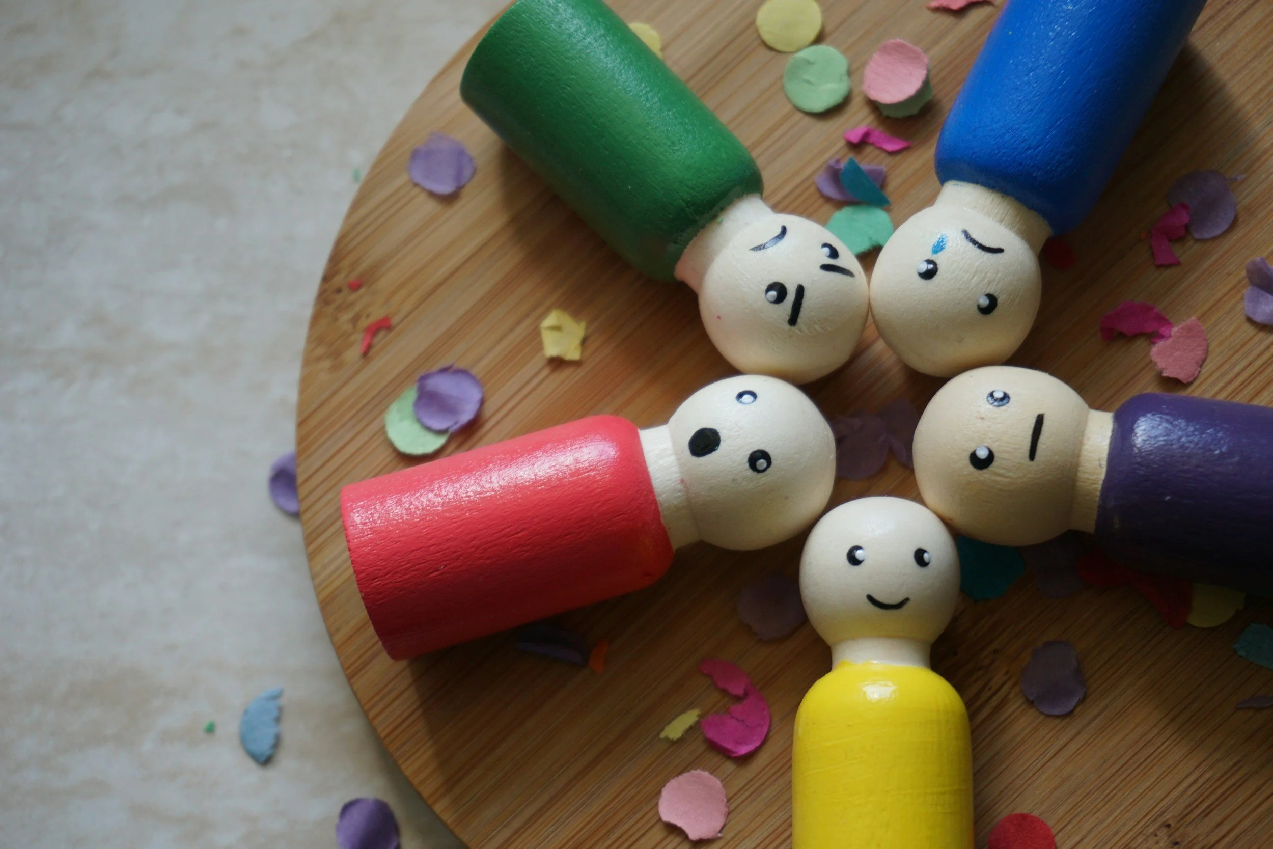 Colorful wooden dolls with painted faces lying on a round wooden surface, surrounded by scattered colorful confetti paper.