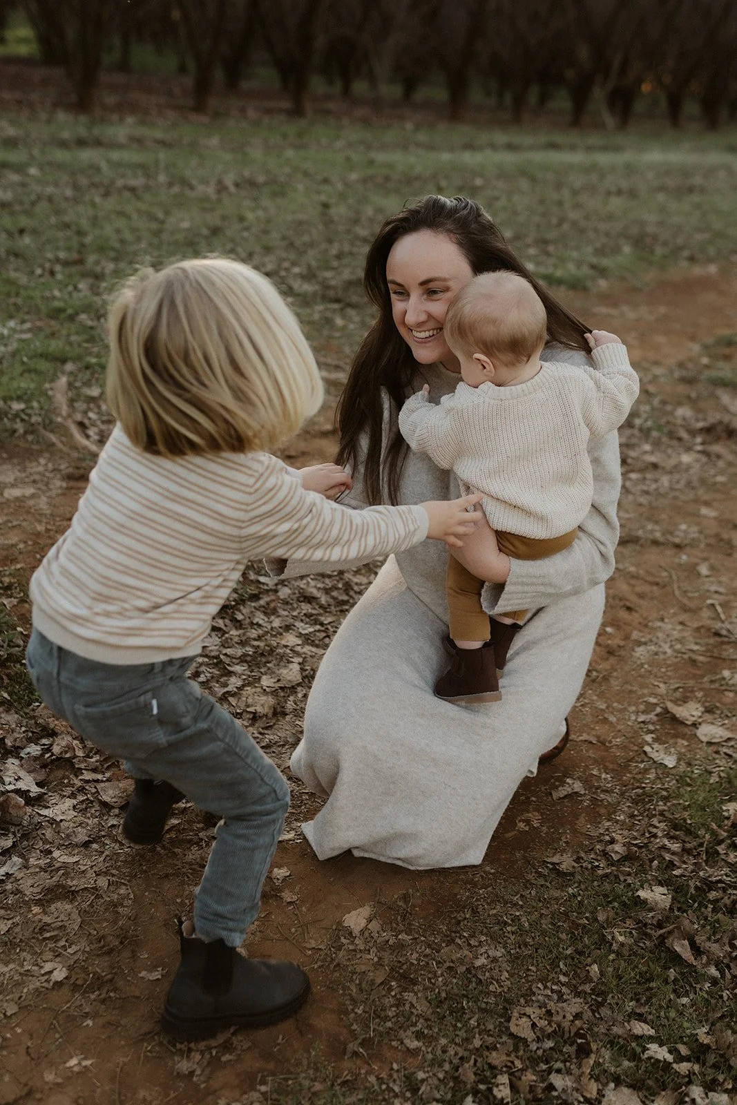 A woman with long dark hair smiling and holding a small child in cream-knit sweater, while another child with blond hair, striped shirt, and gray pants reaches towards the child, outside on a dirt path with brown fallen leaves around, surrounded by trees.