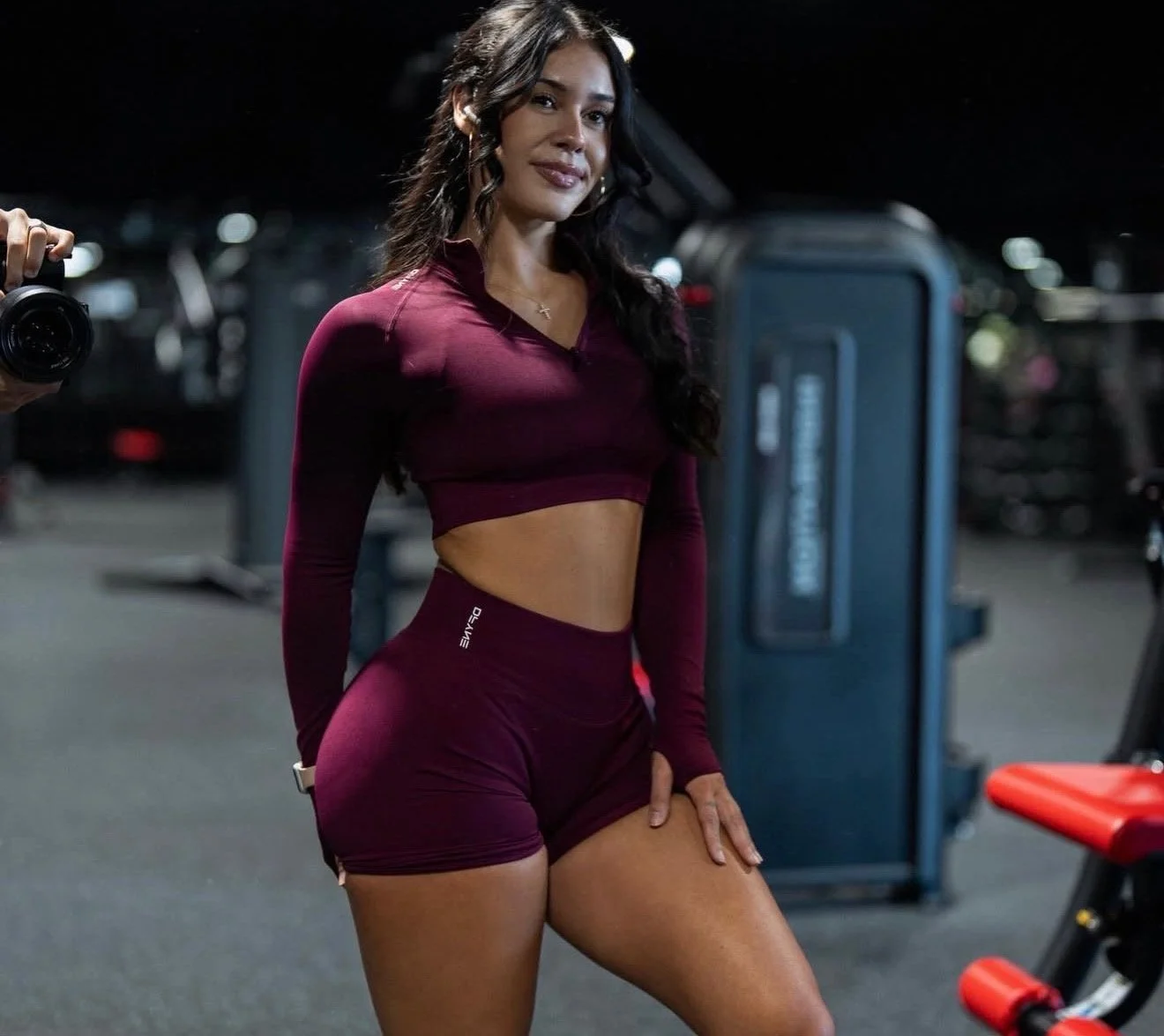 Temecula female athlete in workout attire wearing long-sleeve crop top and shorts, standing in front of gym equipment at Self Made Training Facility—focused on strength training, conditioning, and high-performance fitness.