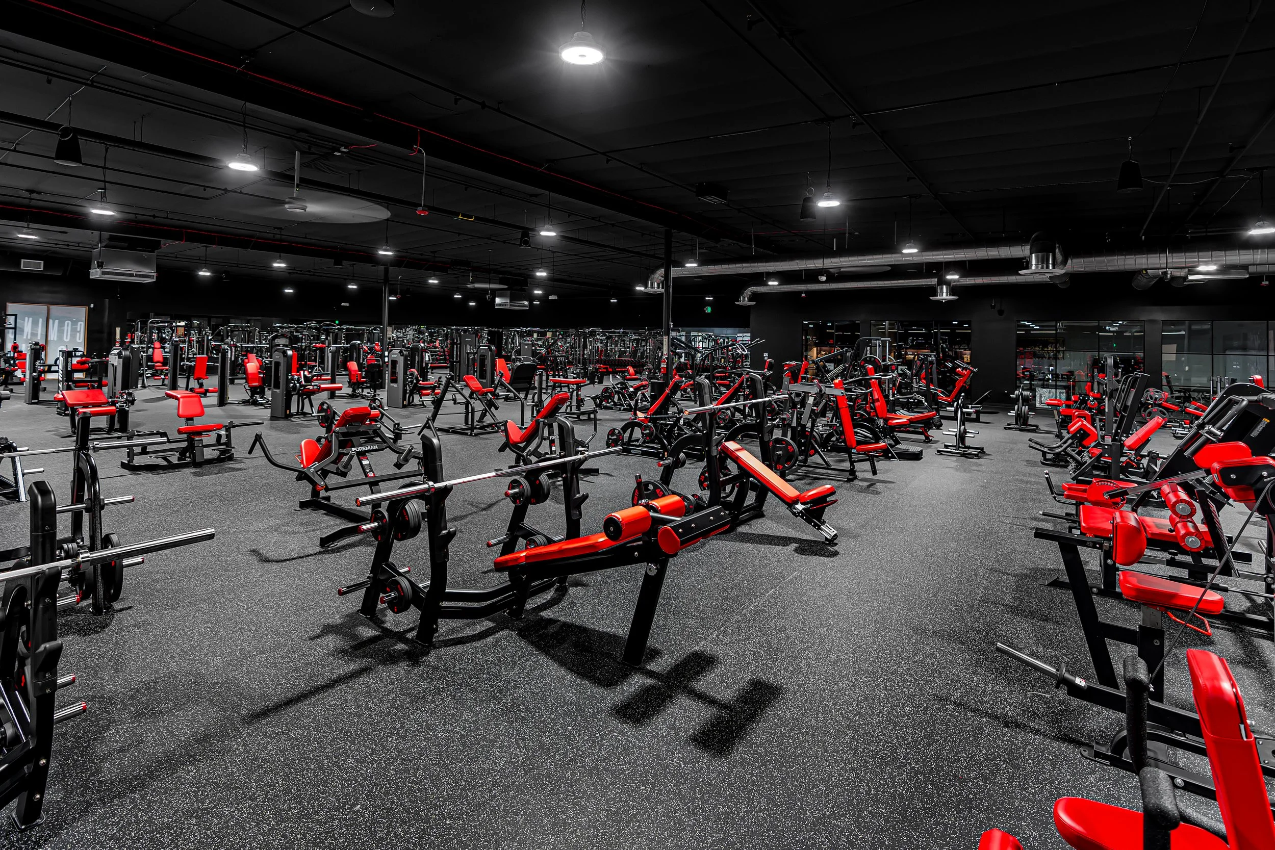 Interior of a gym with various exercise machines and weights, all predominantly black and red, on black rubber flooring.
