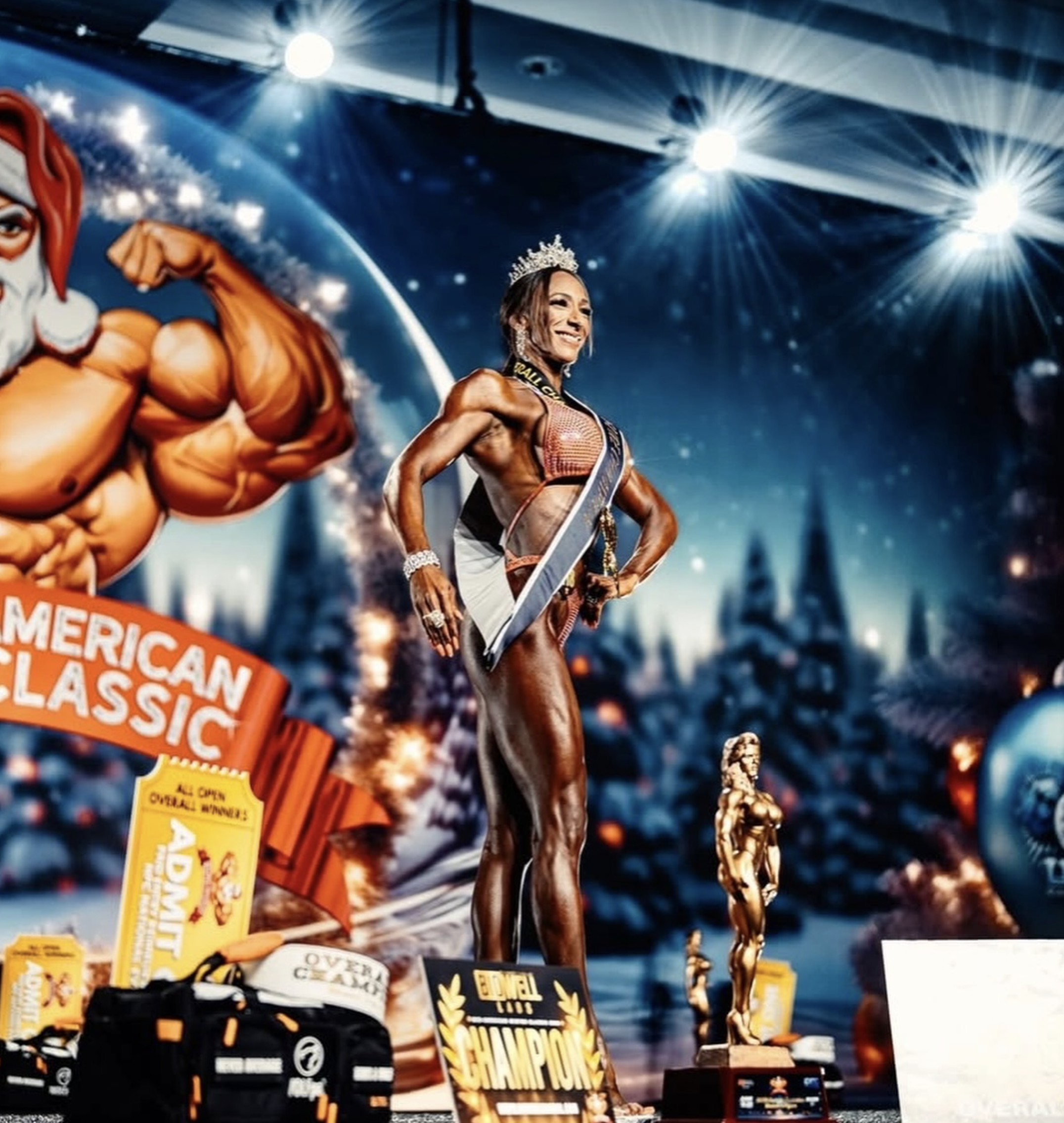 A female bodybuilder wearing a sash, crown, and jewelry on stage at a bodybuilding competition, smiling with her hands on her hips, with a muscular physique. The background features a large muscular cartoon figure, a