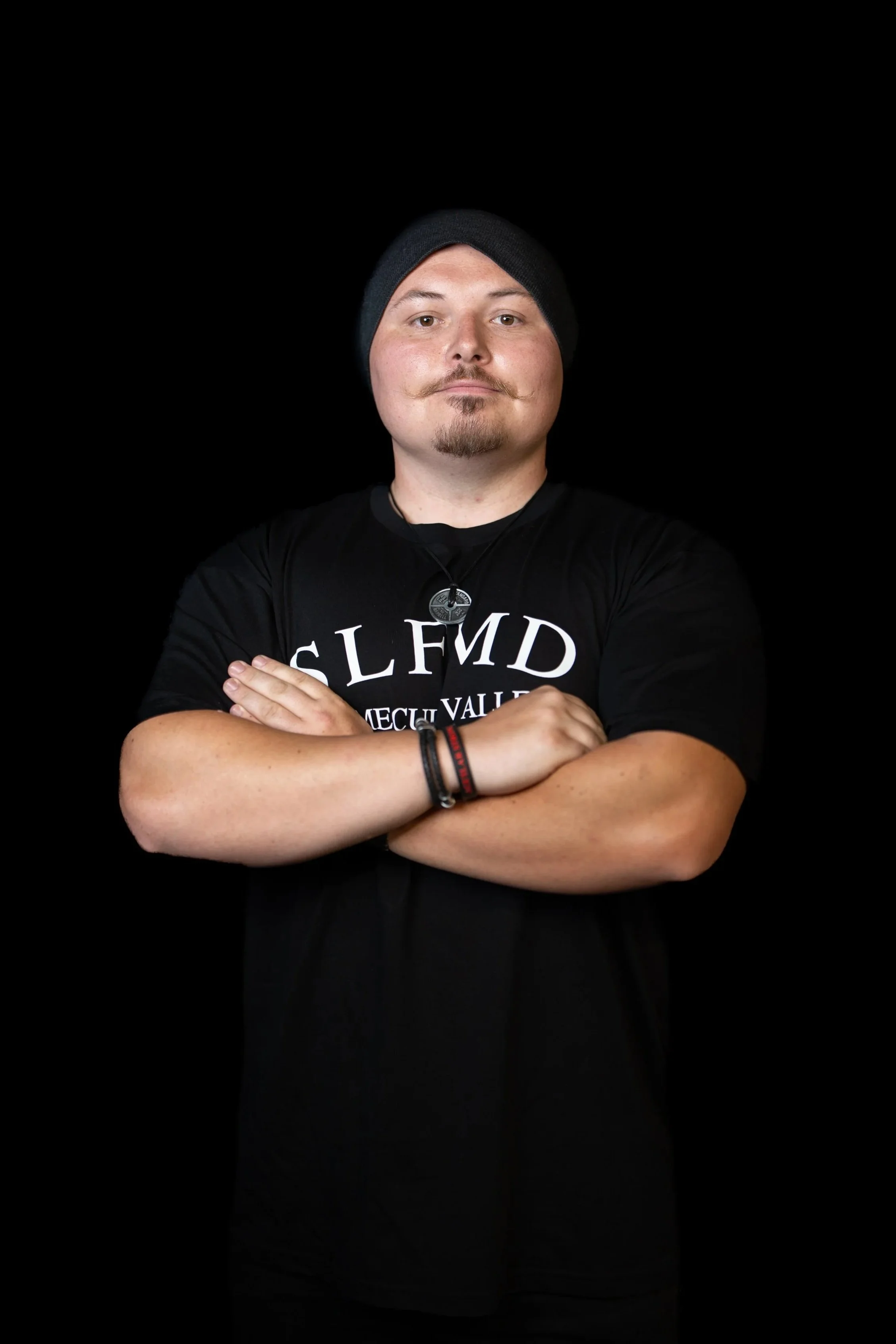 Temecula personal trainer with goatee wearing black beanie and SLFMD shirt, standing with arms crossed against black background at Self Made Training Facility—specializing in strength training, fat loss, and coaching clients on the autism spectrum.