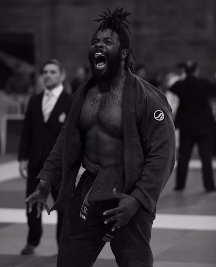 Man with dreadlocks, shirtless and wearing a martial arts gi jacket, shouting and celebrating in a martial arts dojo.