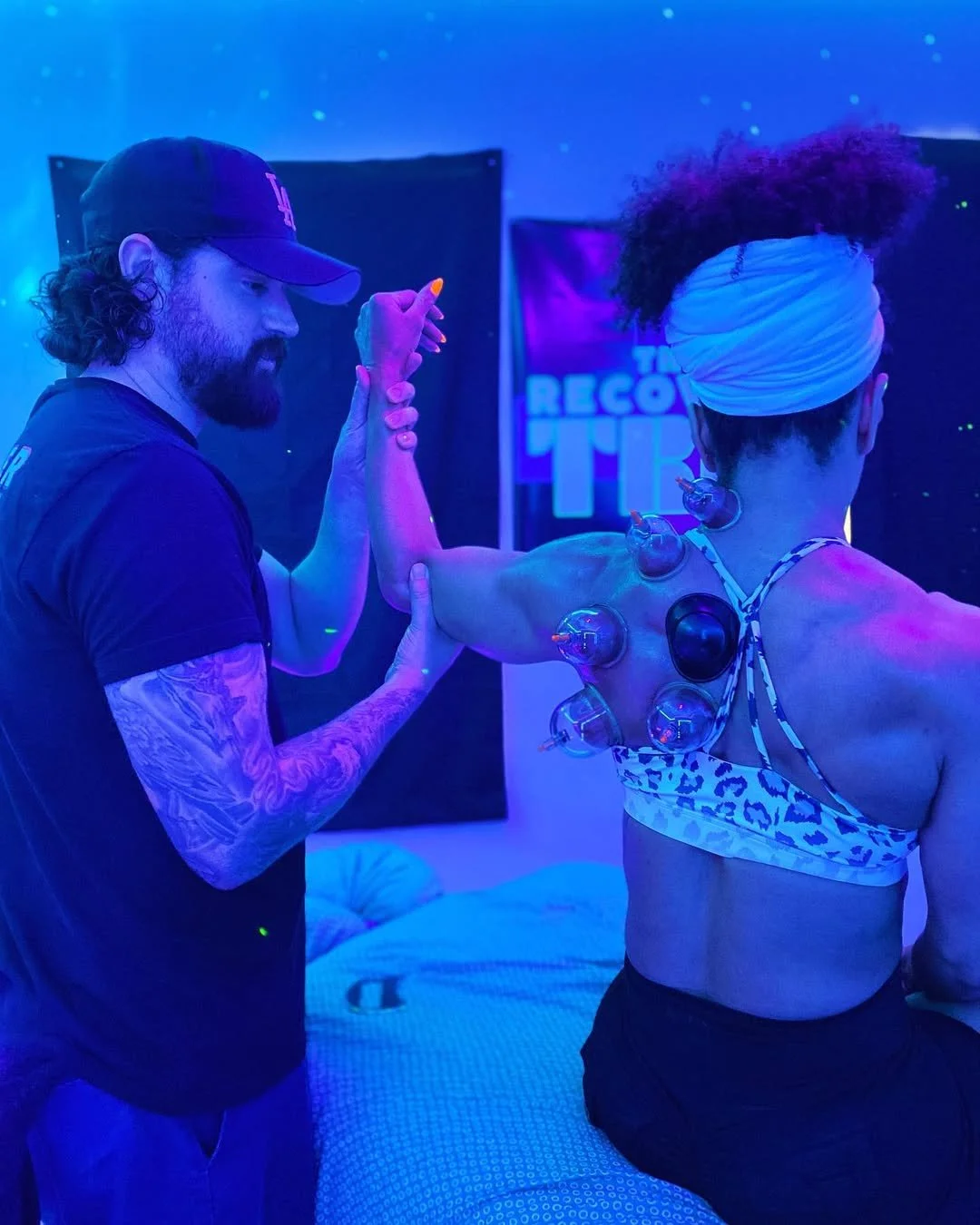 Man with tattoos and beard performing sports massage recovery session assisting female client with dynamic movement while cupping therapy is applied under black lights.-- sports recovery massage therapist injury prevention muscle recovery