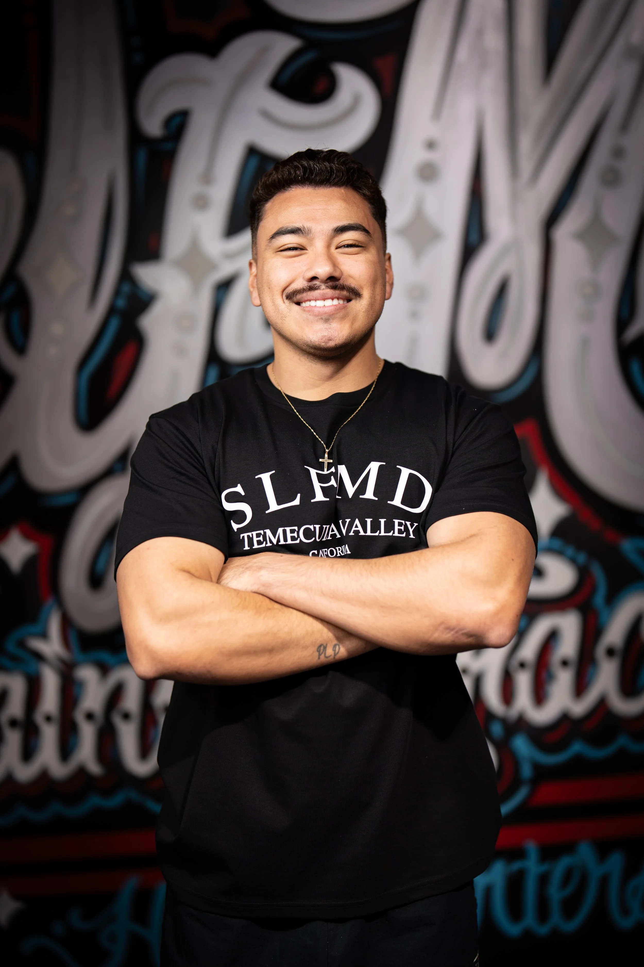 Smiling Temecula personal trainer with short dark hair and goatee wearing black SLFMD shirt and gold necklace, standing with arms crossed at Self Made Training Facility—specializing in strength training and personalized fitness coaching.