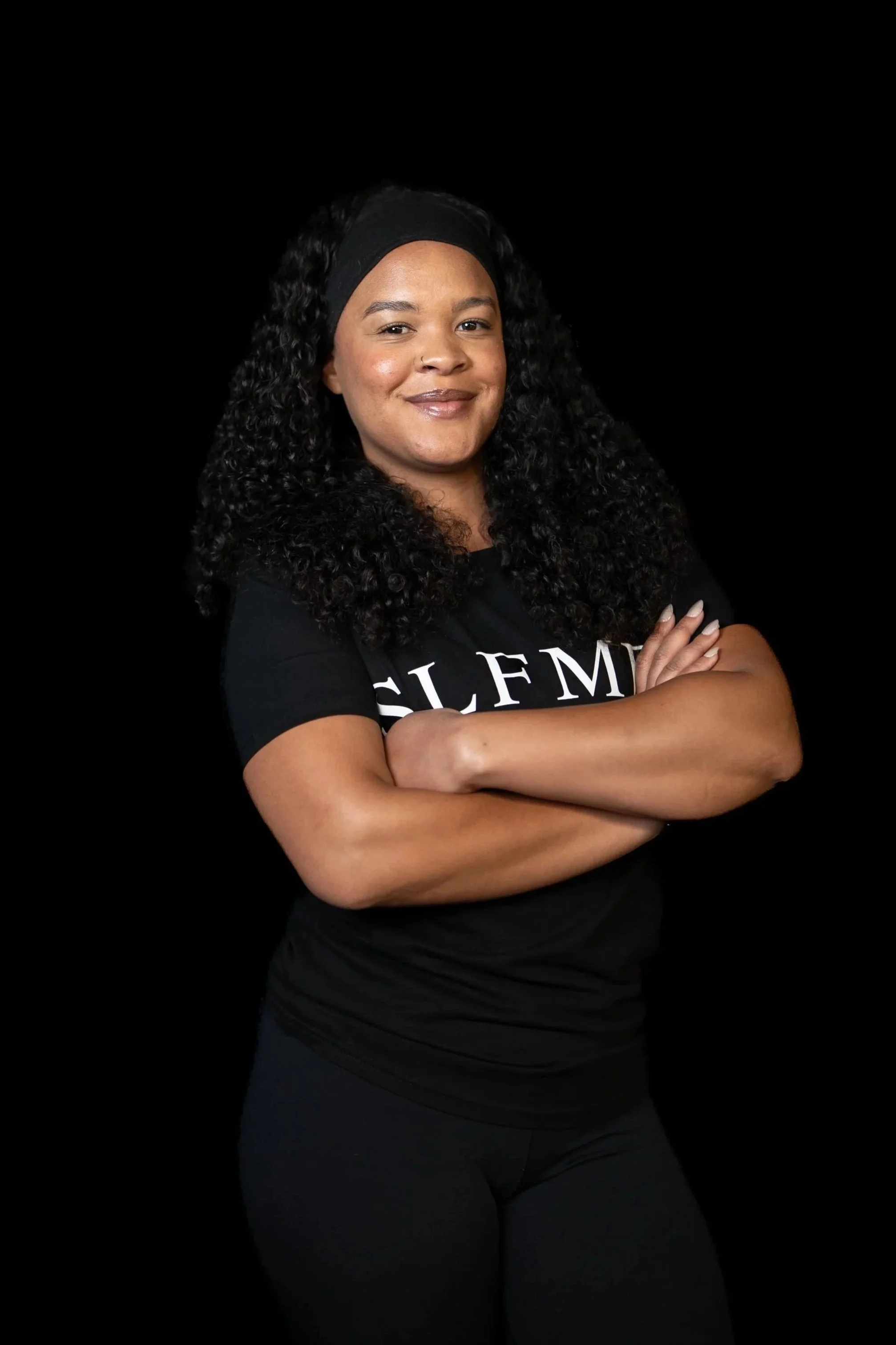 Temecula personal trainer with curly black hair wearing a black headband and SLFMD shirt, confidently standing with arms crossed against a black background at Self Made Training Facility—specializing in strength training, and weight loss.