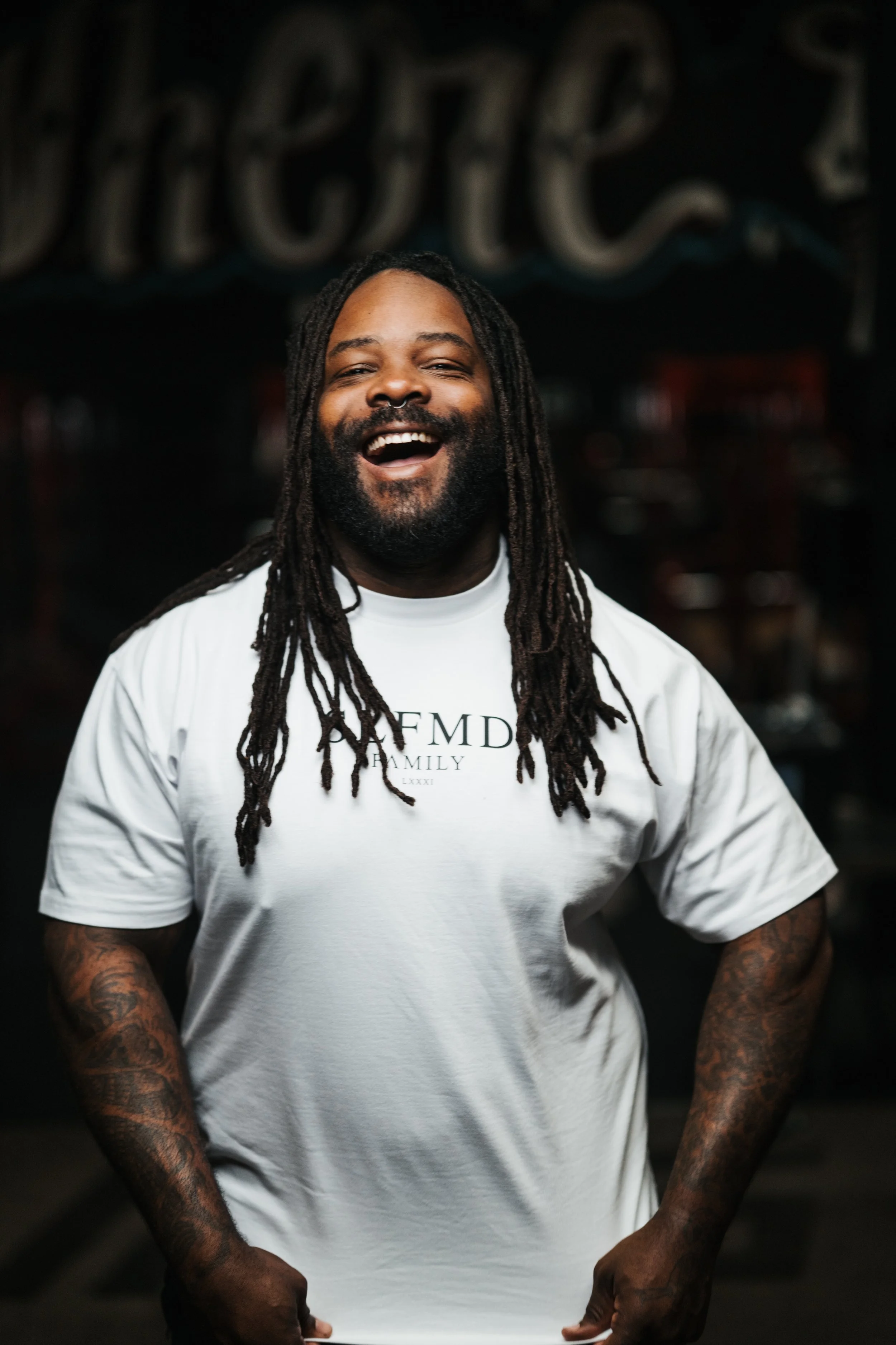 A man with long dreadlocks and tattoos on his arms, smiling widely while wearing a white T-shirt with a logo.