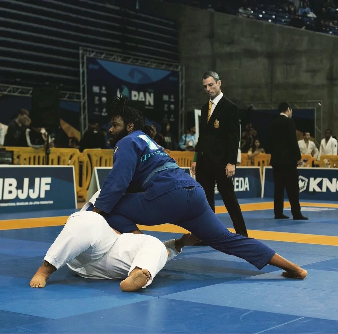 Two judo competitors in blue and white gi on the mat during a match, with a referee in a black suit observing nearby, in an indoor sports arena with spectators and banners in the background.