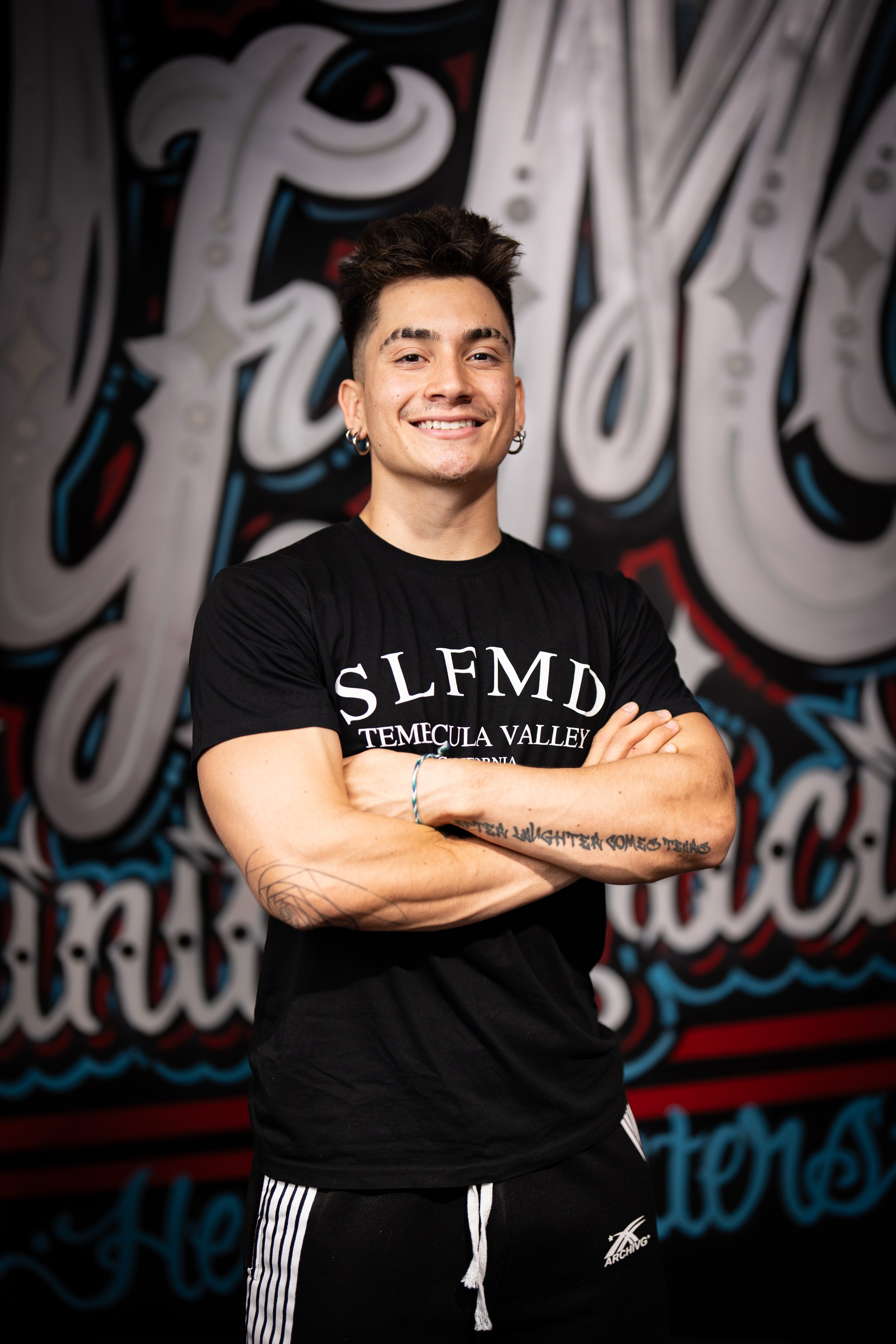 A young man with dark hair and earrings smiling with arms crossed, standing in front of a colorful graffiti-style mural.