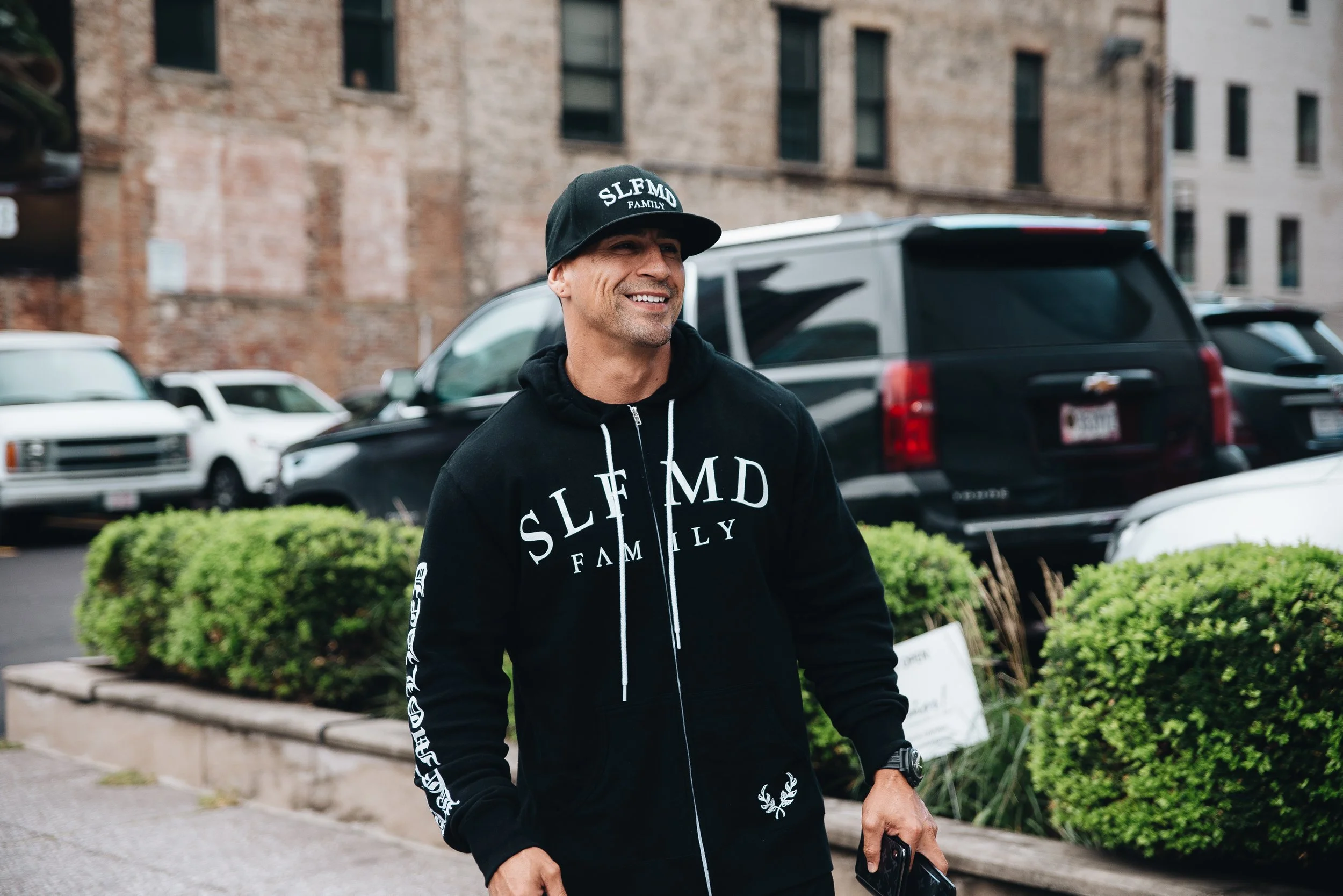 A smiling man wearing a black hoodie and cap, both with white text, walking outdoors in a parking lot with parked cars and brick buildings in the background.