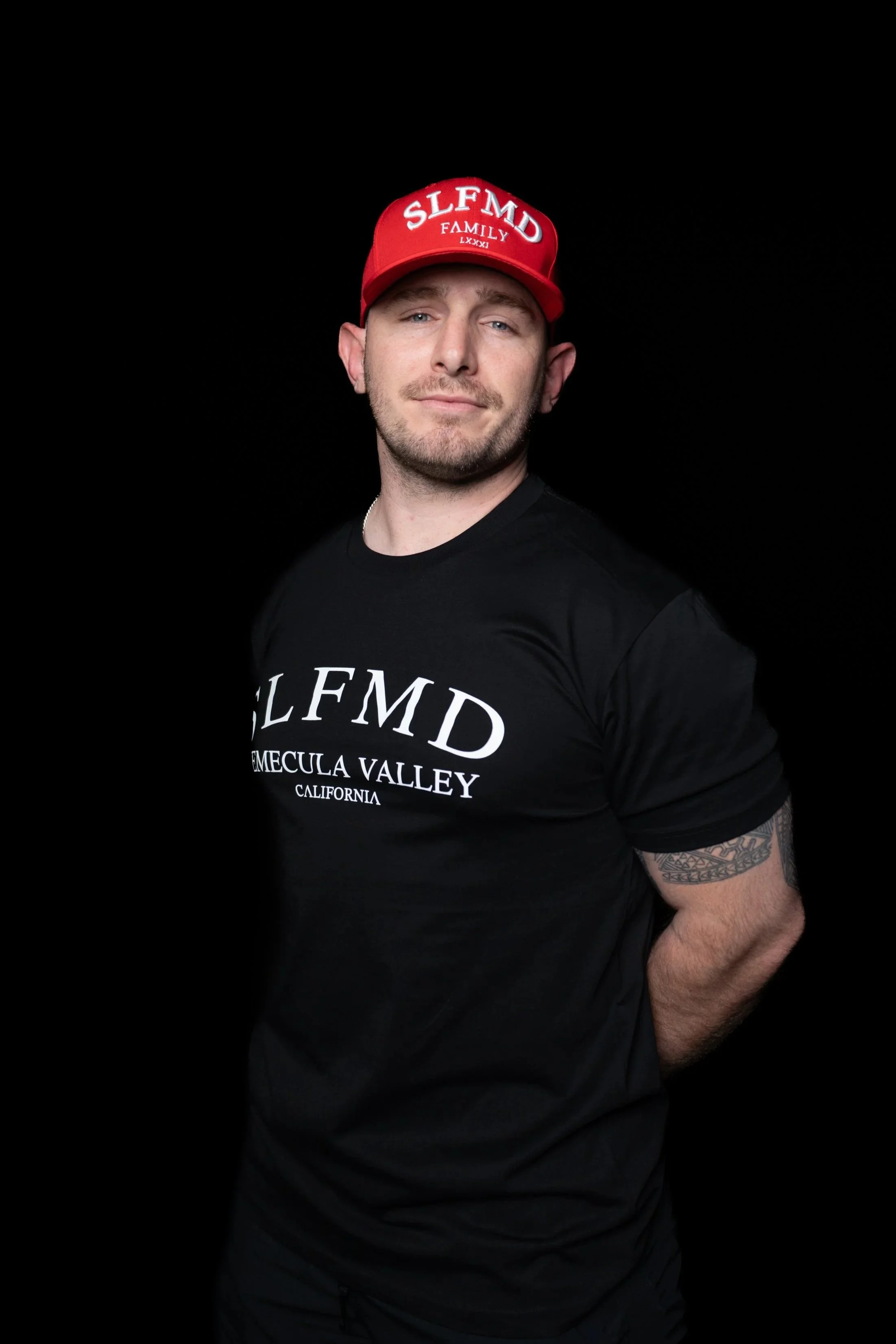 Temecula personal trainer wearing SLFMD Temecula Valley shirt and red baseball cap, standing against a black background at Self Made Training Facility—specializing in strength training, fat loss, and athlete development.