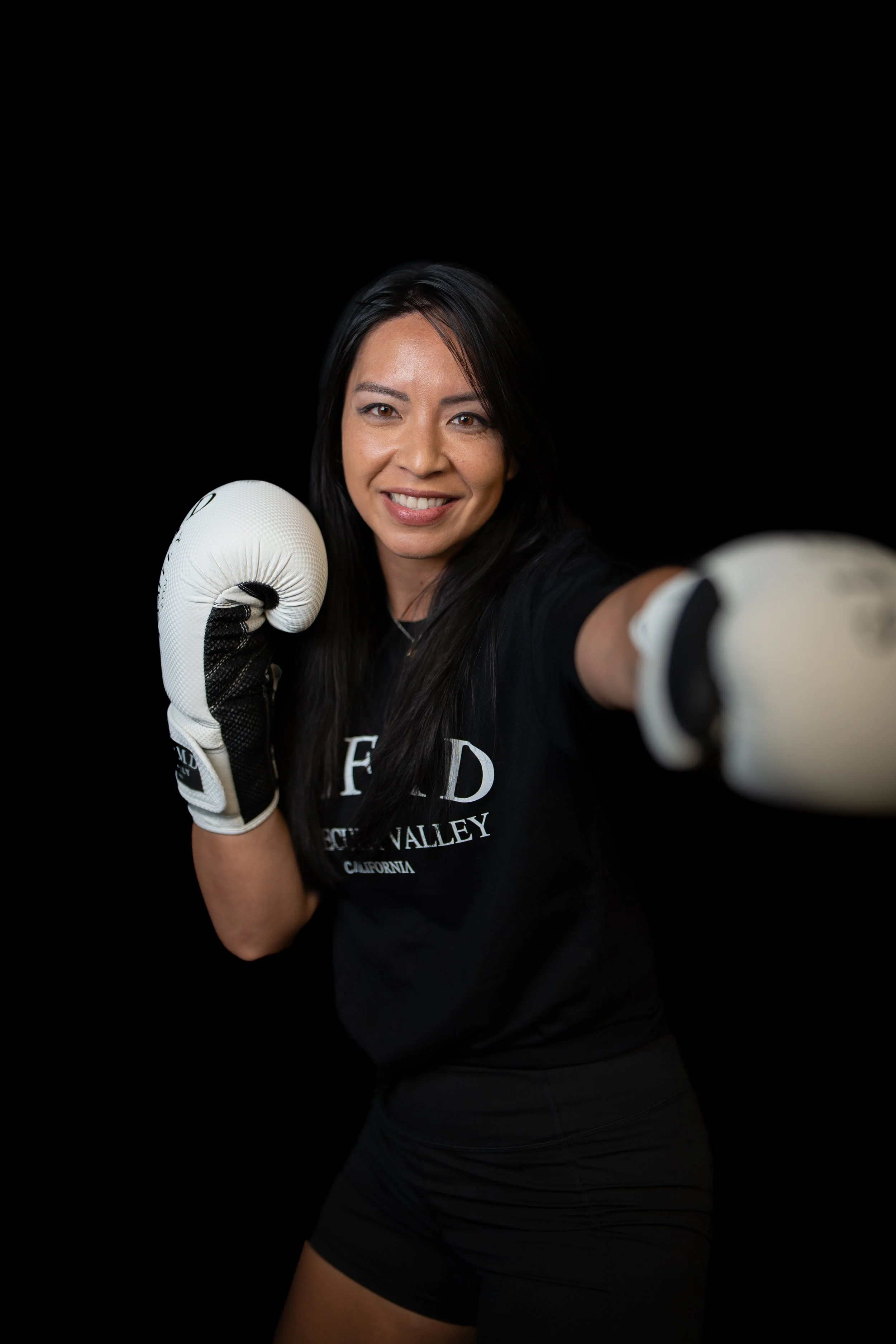 Temecula female boxing coach in black athletic wear and gloves smiling in fighting stance against black background at Self Made Training Facility—specializing in boxing, conditioning, and women’s athletic training.