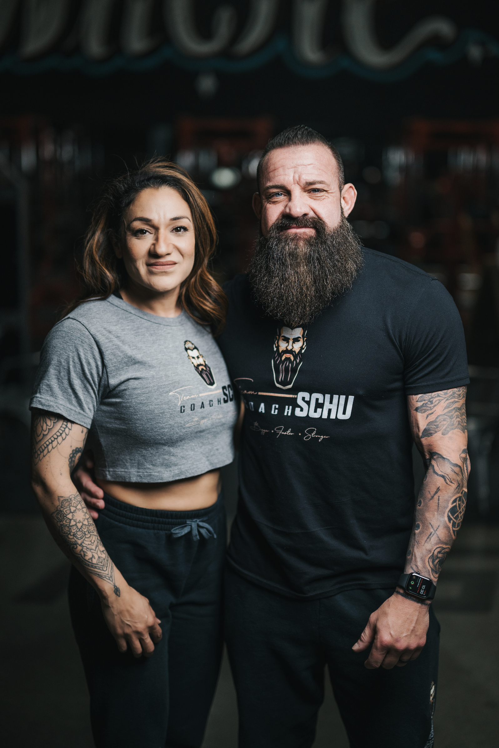 A woman and a man standing together in a gym. The woman has medium-length brown hair, tattoos on her left arm, and is wearing a gray cropped t-shirt and black pants. The man has a long beard, tattoos on both arms, and is wearing a black T-shirt and a smartwatch. They are both smiling slightly and posing for the camera.