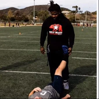 A woman in a black hoodie walking on a sports field, looking down at a child lying on the ground.