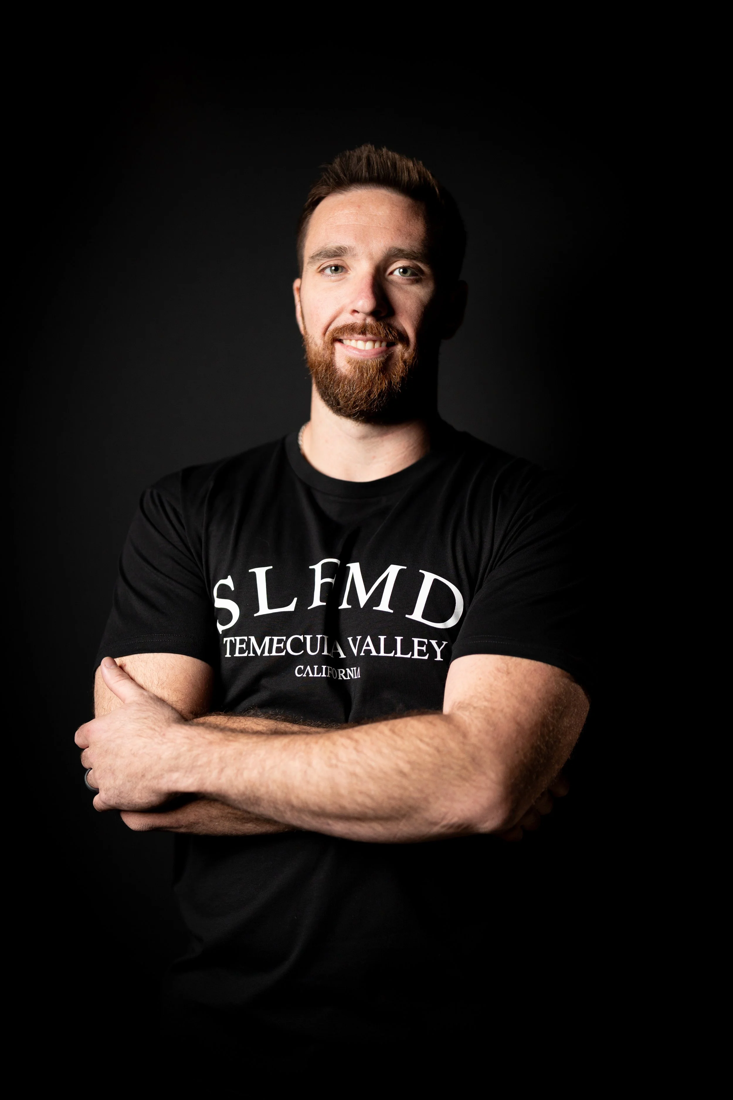 Smiling Temecula personal trainer with beard and short hair wearing black SLFMD shirt, standing with arms crossed against dark background at Self Made Training Facility—focused on client transformation, strength training, and fat loss.