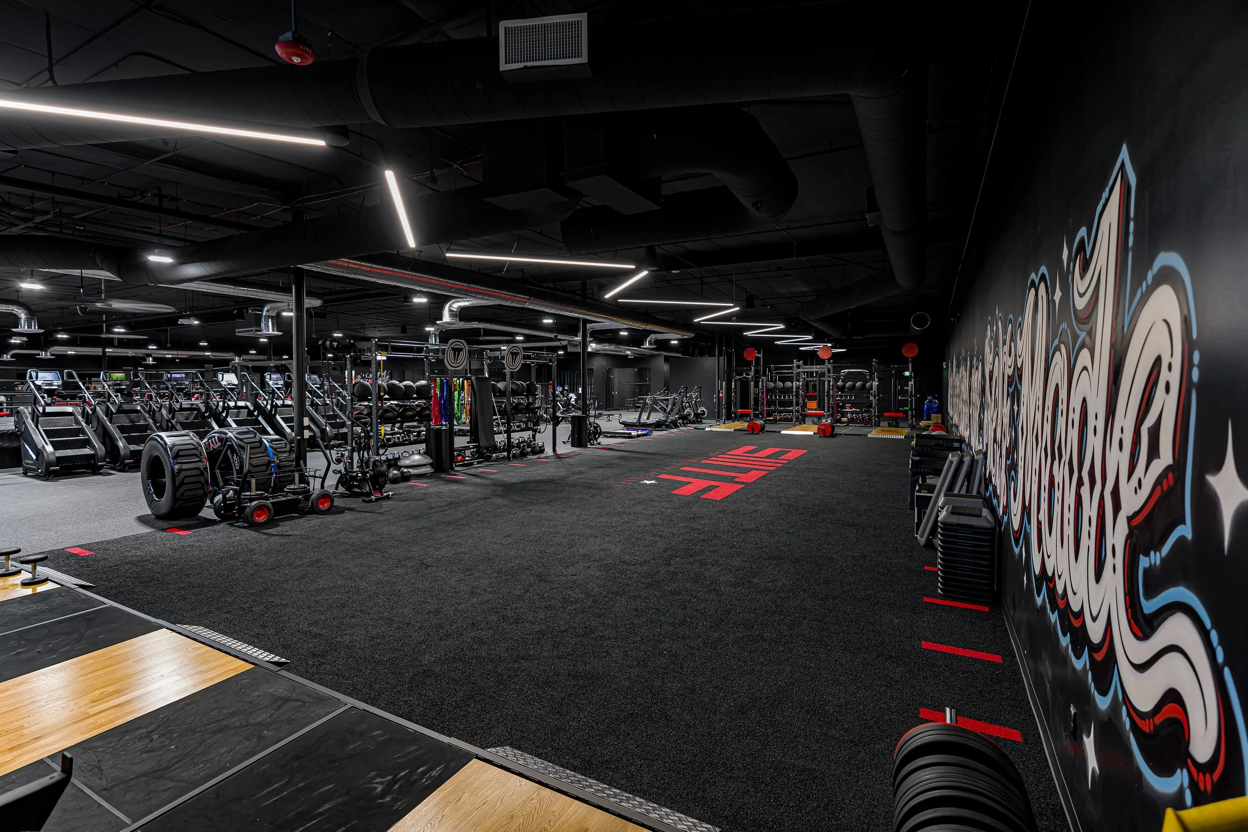 Temecula 24/7 gym with weightlifting and cardio equipment, graffiti art on black walls, and bright overhead lighting at Self Made Training Facility—elite training space for athletes, bodybuilders, and personal trainers.
