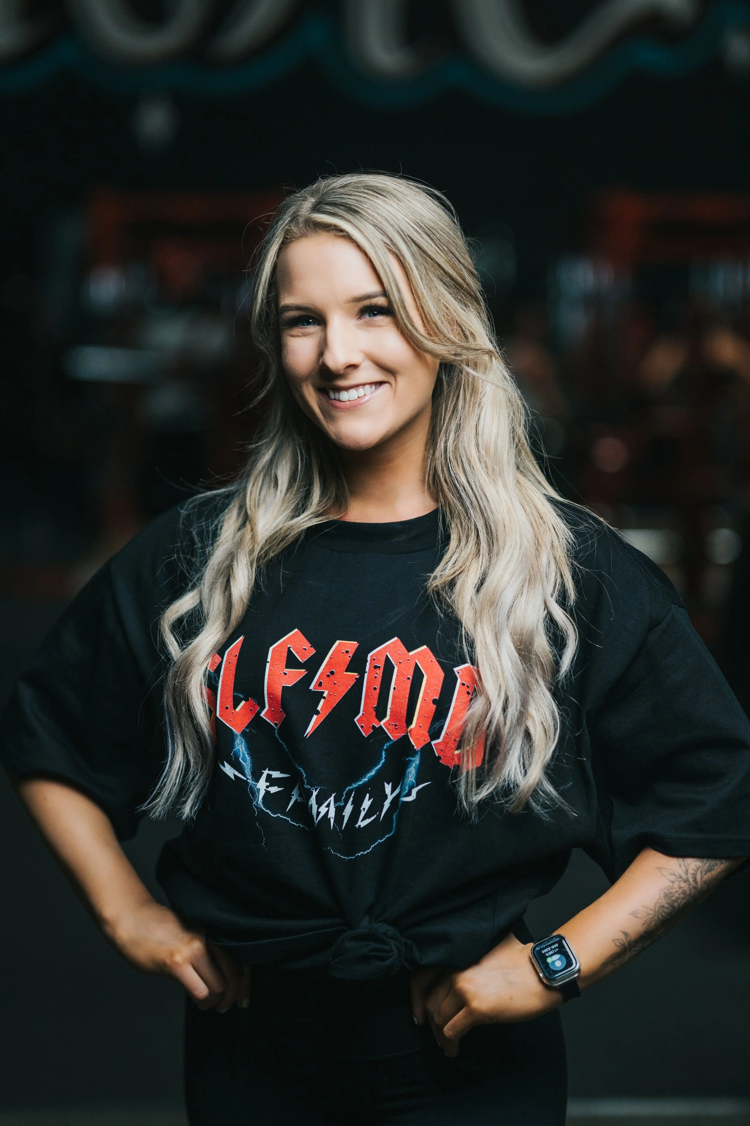 Smiling Temecula personal trainer with long blonde hair in a black SLFMD shirt, standing hands on hips at Self Made Training Facility—focused on women’s fitness and strength training.