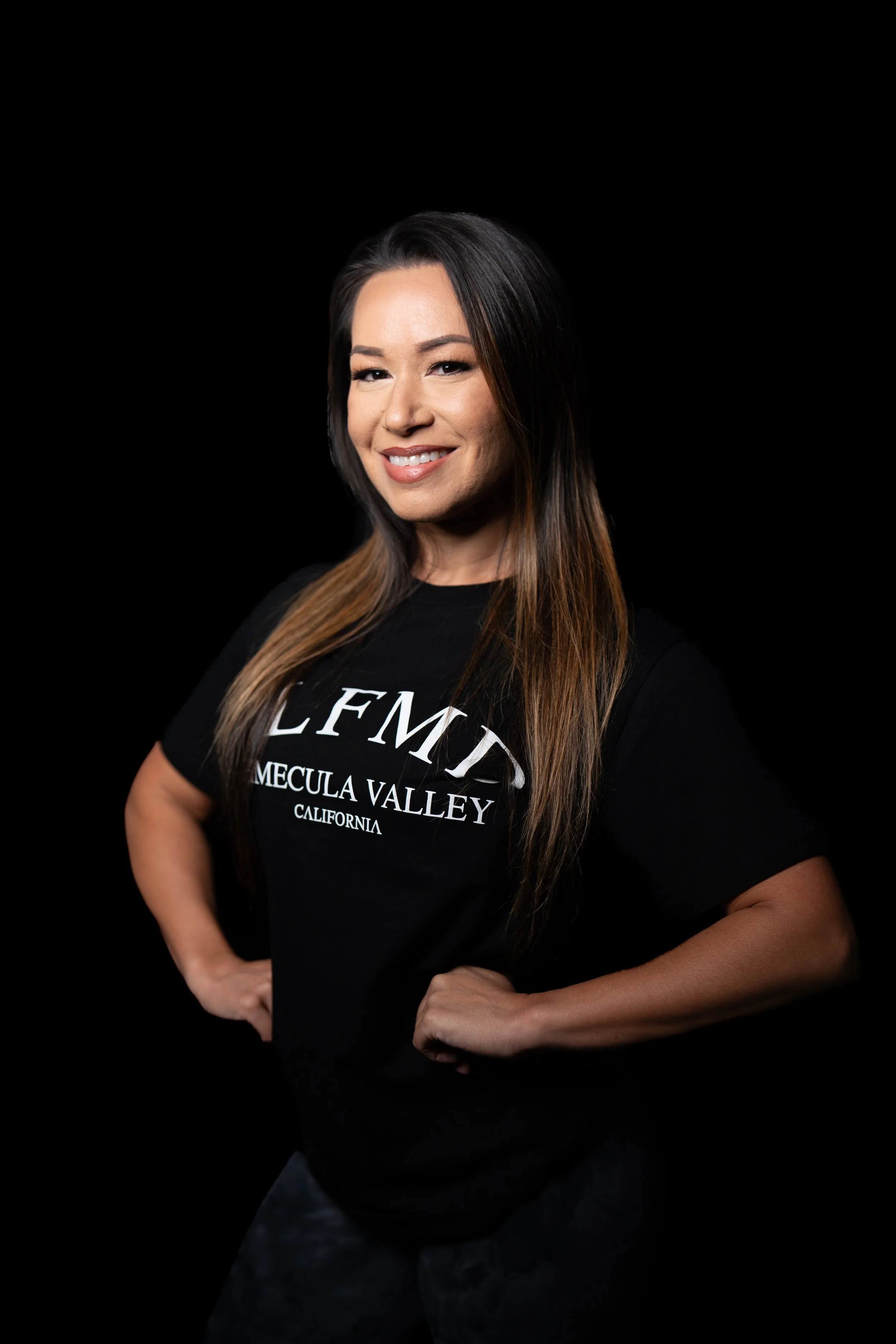 Smiling Temecula personal trainer with long brown hair wearing black SLFMD shirt, standing with hands on hips against a black background at Self Made Training Facility—focused on women’s strength training and fitness coaching.