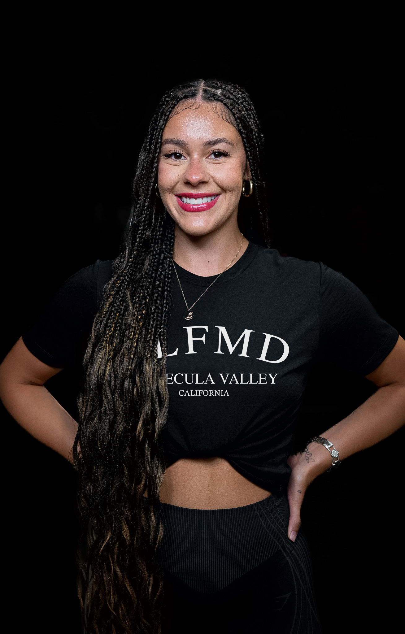 Smiling Temecula personal trainer and women’s life coach with long braided hair wearing black SLFMD crop top, standing against dark background at Self Made Training Facility—focused on holistic fitness, mindset, and strength training for women.