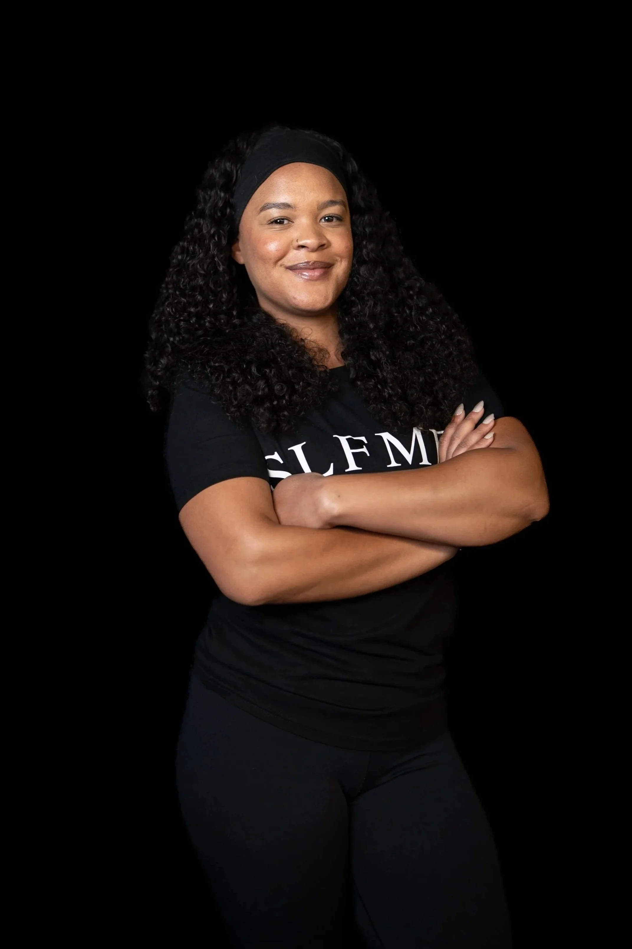 Temecula personal trainer with curly hair wearing a black SLFMD shirt and headband, standing with arms crossed against a black background at Self Made Training Facility—focused on strength training and women’s fitness.