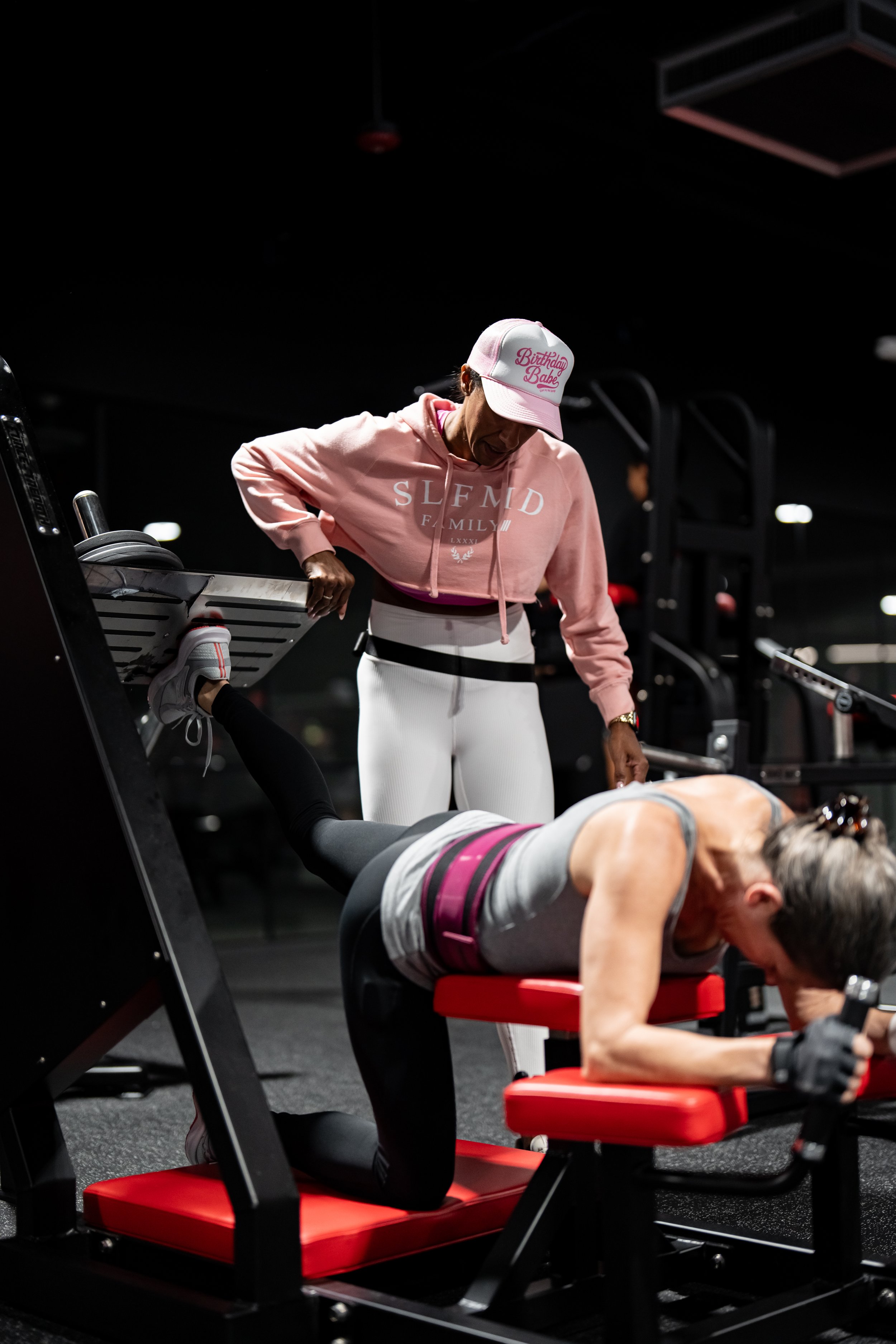 Female athlete using a leg curl machine with assistance from a personal trainer in a pink hoodie at Self Made Training Facility Temecula Valley, highlighting expert coaching and strength training in a high-performance gym setting.