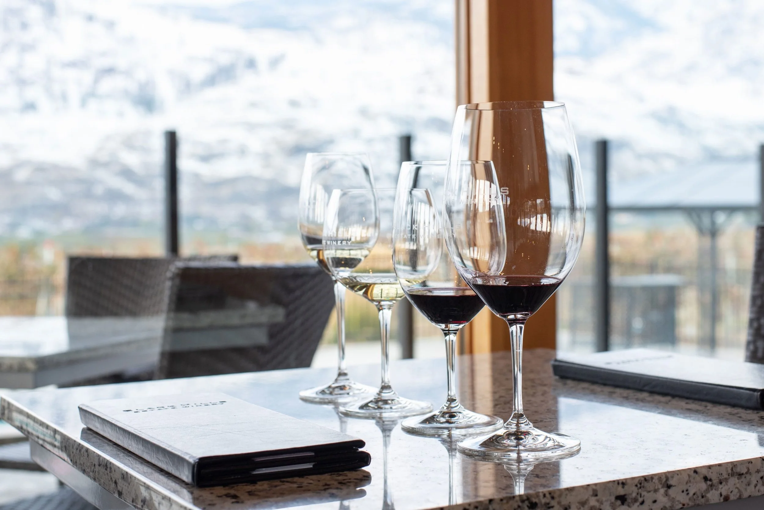 Four wine glasses with varying amounts of white and red wine on a table in a bright room with mountains and snow outside.