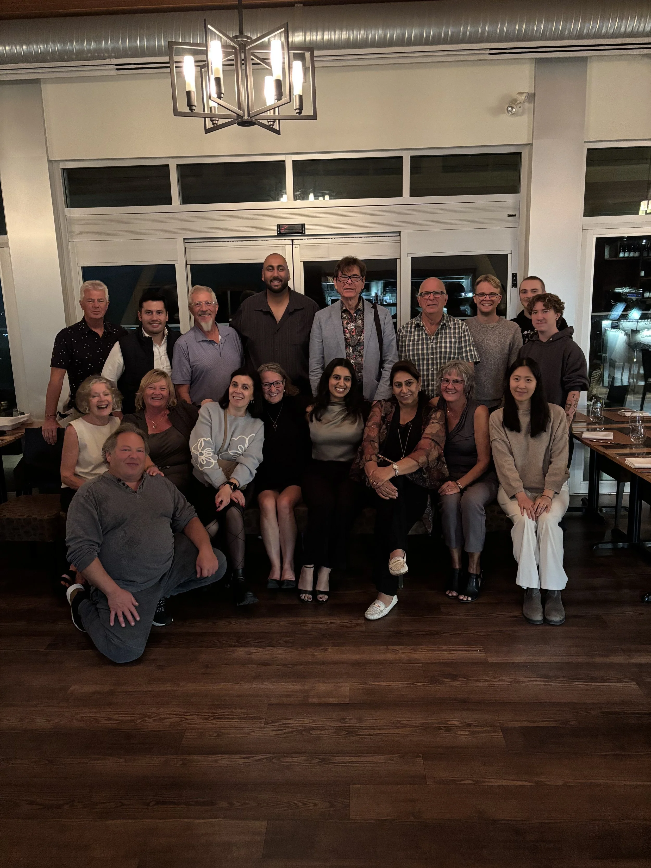 Group of people posing for a photo in a restaurant dining area with wooden floors, a modern chandelier, and a light-colored wall with large windows.