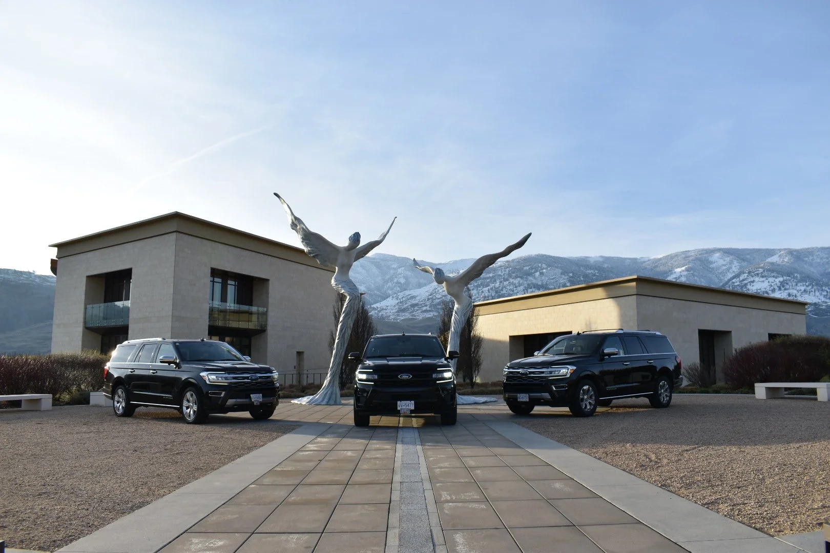 Three black SUVs parked in front of modern buildings with large abstract metallic sculptures of two women with outstretched arms and legs, set against a mountainous landscape with snow-capped peaks.
