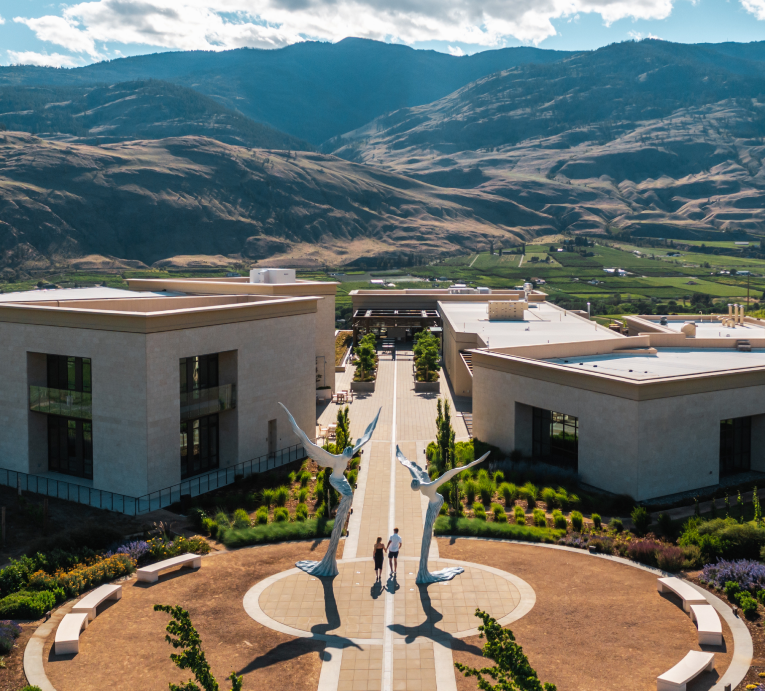 Aerial view of a modern building complex with sculpture of two birds and two people walking on a pathway, mountains in the background.