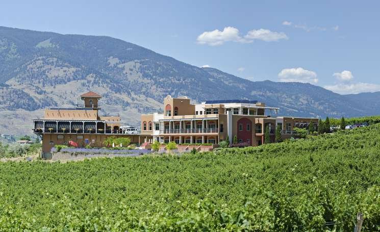 Luxury building complex with multiple levels and balconies, set against a lush green vineyard with mountains in the background under a partly cloudy sky.