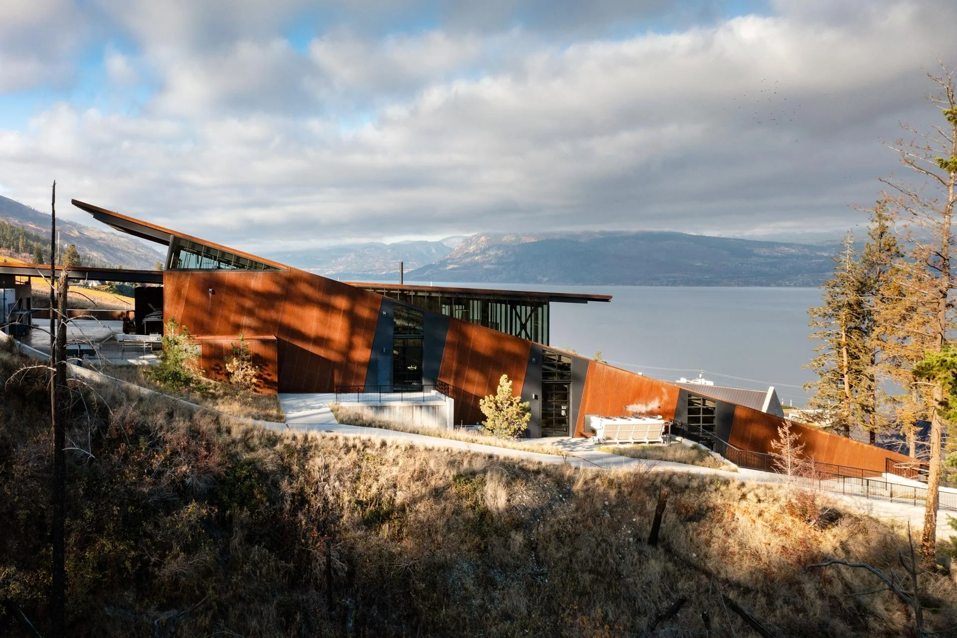 A modern building with angular, rust-colored metal and glass walls situated on a hillside overlooking a body of water with mountains in the distance. The scene includes dry grass and trees with some leaves turning color.