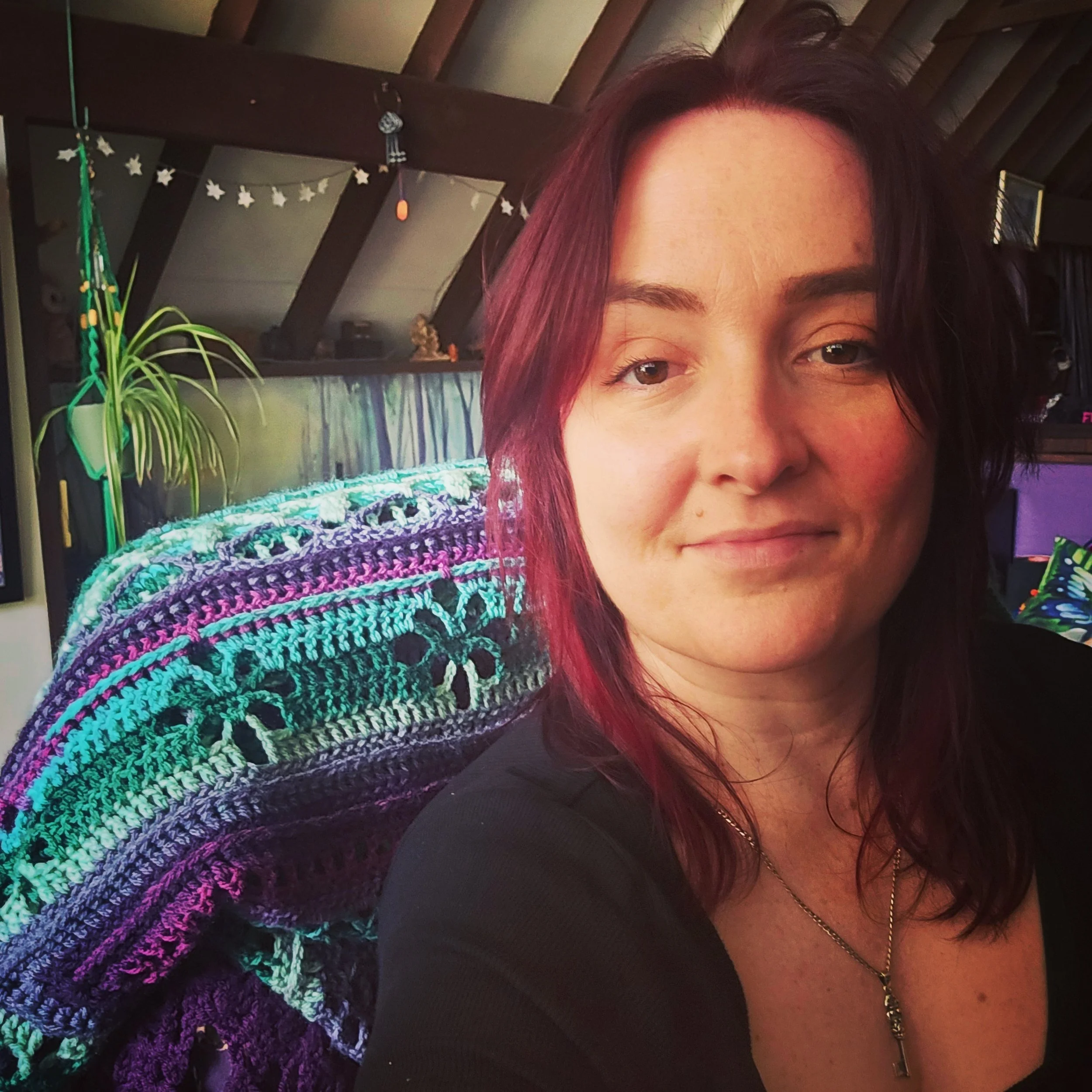 A woman with shoulder-length dark red hair, wearing a black top and a necklace, smiling softly in a cozy, decorated room with a colorful crocheted blanket, houseplants, and decorative items.