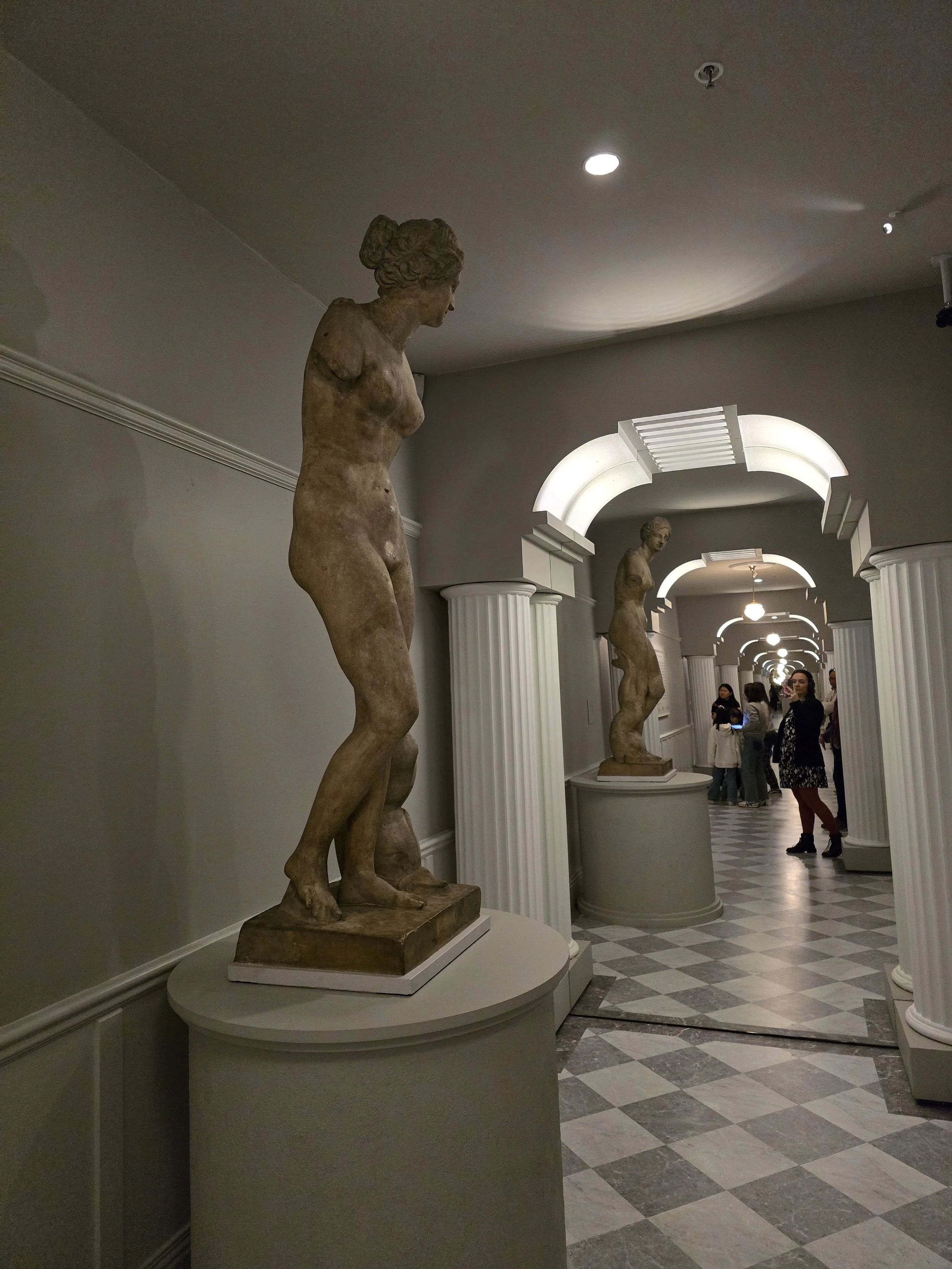 Museum hallway with classical statues on pedestals, featuring columns and arches, and visitors in the background.