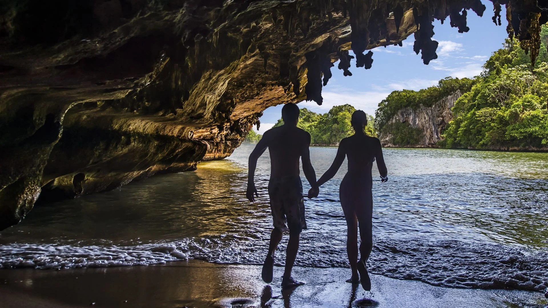A couple holding hands walks along a sandy beach near a rocky cave with lush green trees and a blue sky in the background.