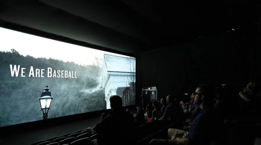 People seated inside a dark room watching a large screen with the words 'We Are Baseball' displayed.