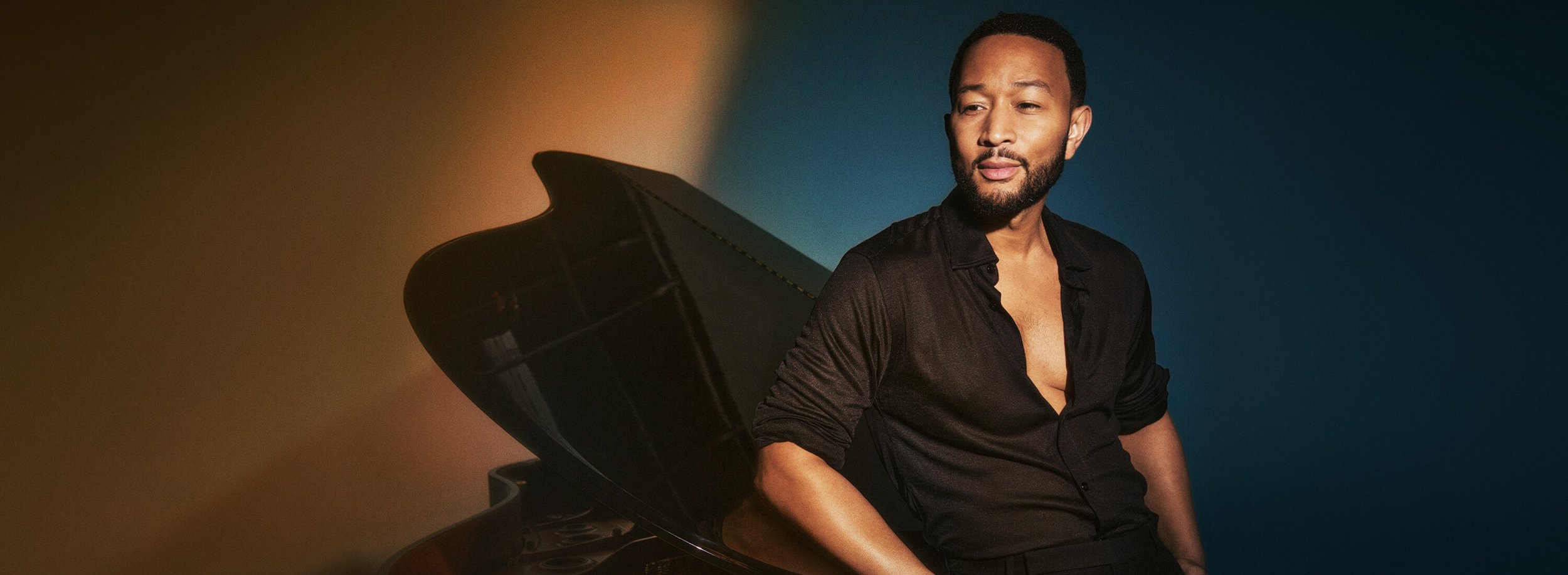 A man with a beard and short hair sitting next to a grand piano, wearing a black partially unbuttoned shirt, with a gradient background from brown to blue.
