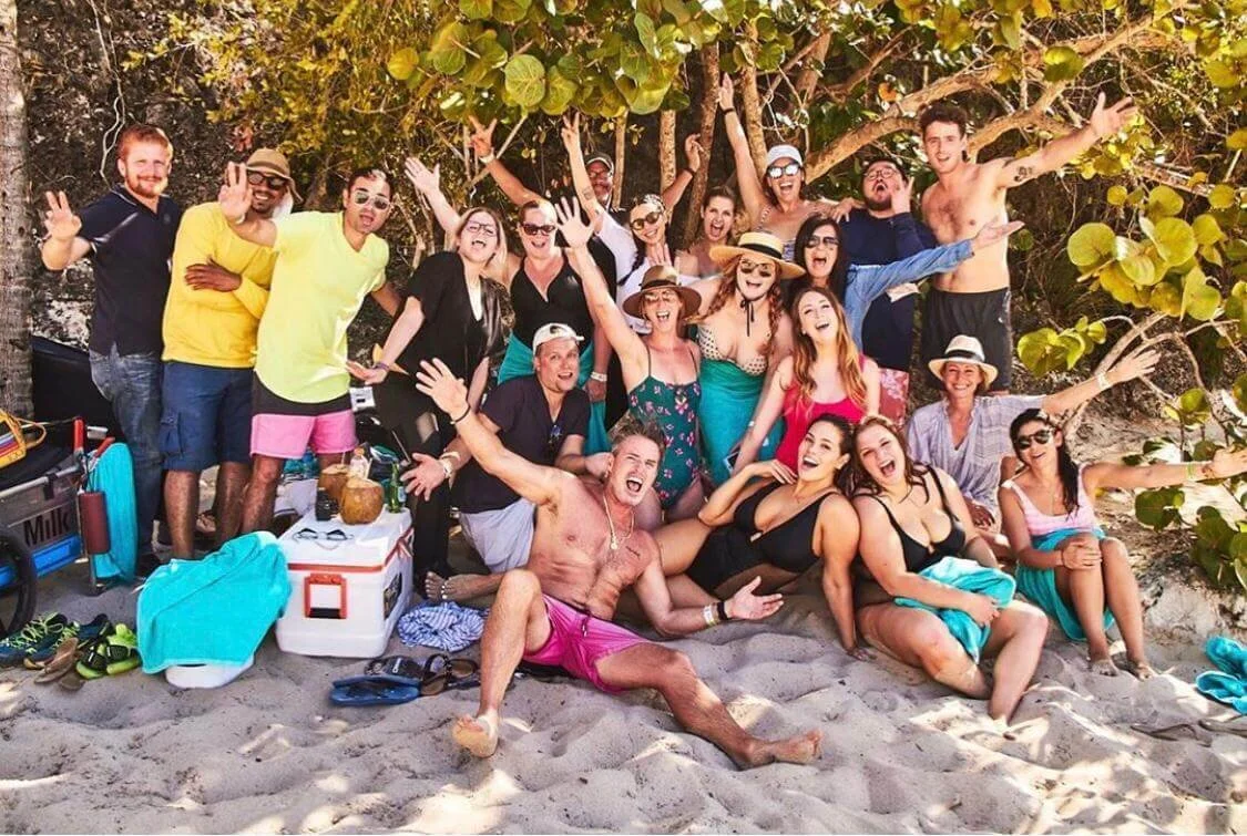 Group of friends posing on the beach, sitting and standing under a tree, enjoying a sunny day with beach gear around them.