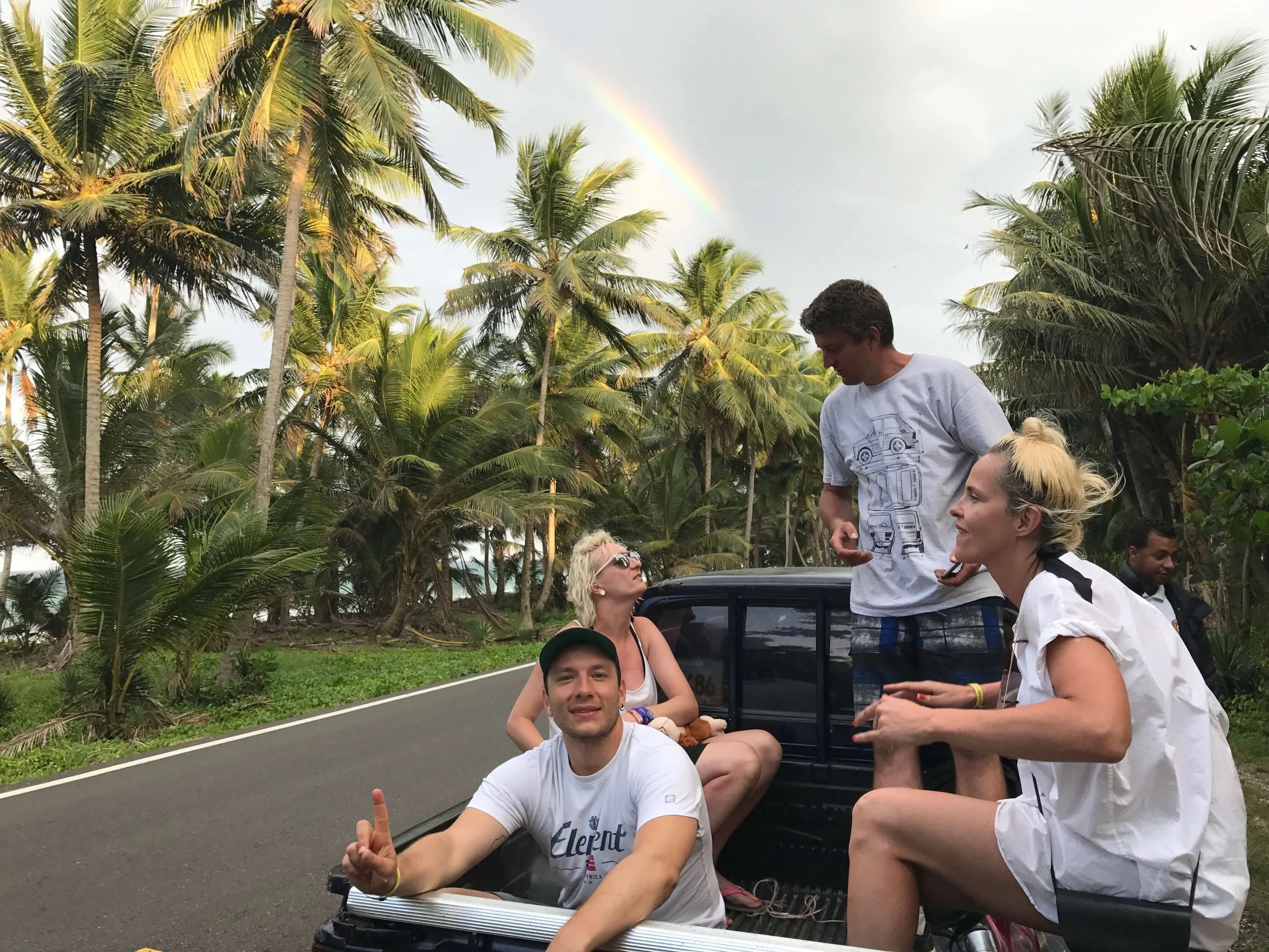 Group of five people, four women and one man, sitting and standing around a small vehicle on a road lined with palm trees, with a rainbow in the sky overhead, in a tropical setting.