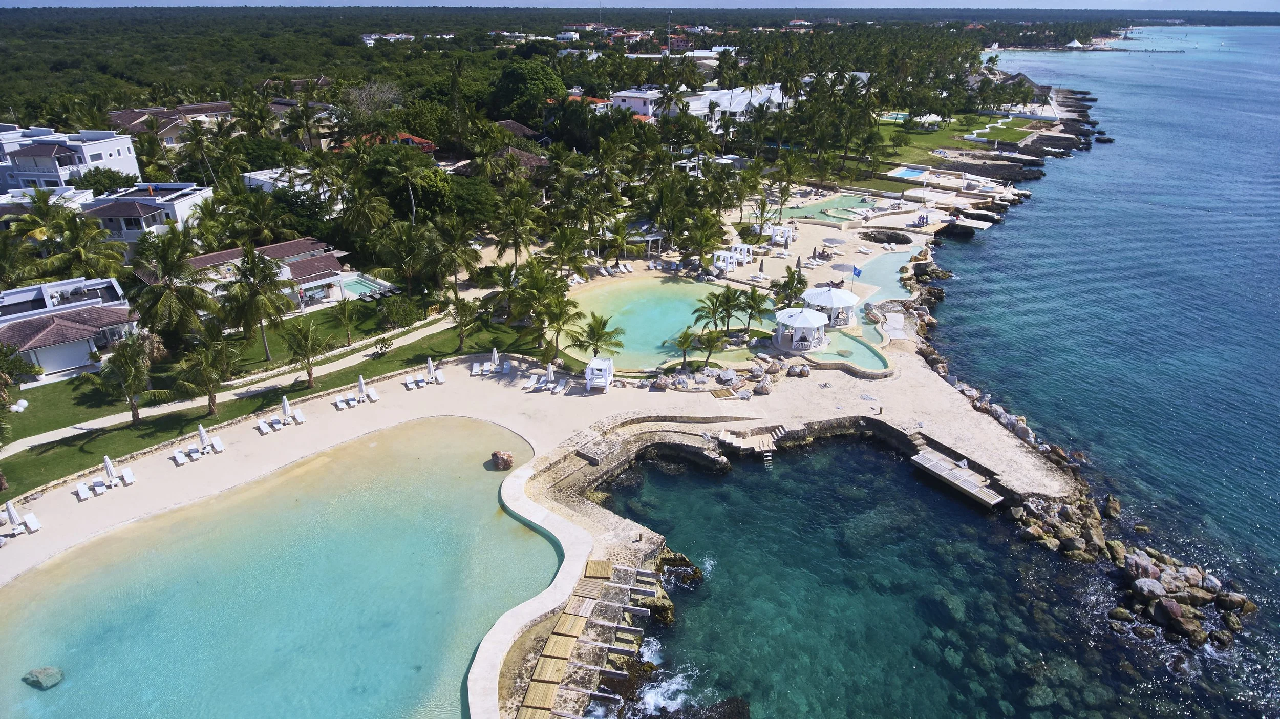 Aerial view of a tropical resort with multiple swimming pools, palm trees, beachfront lounge chairs, and villas along the coast with clear blue water.