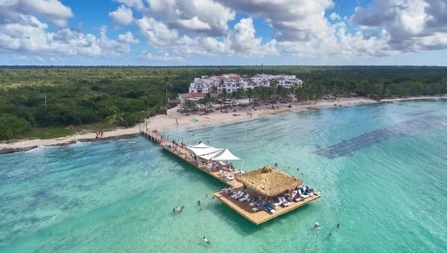 Overhead view of a tropical beach with a pier leading to a thatched-roof hut, anchored boats, and a resort with white buildings surrounded by greenery under a partly cloudy sky.