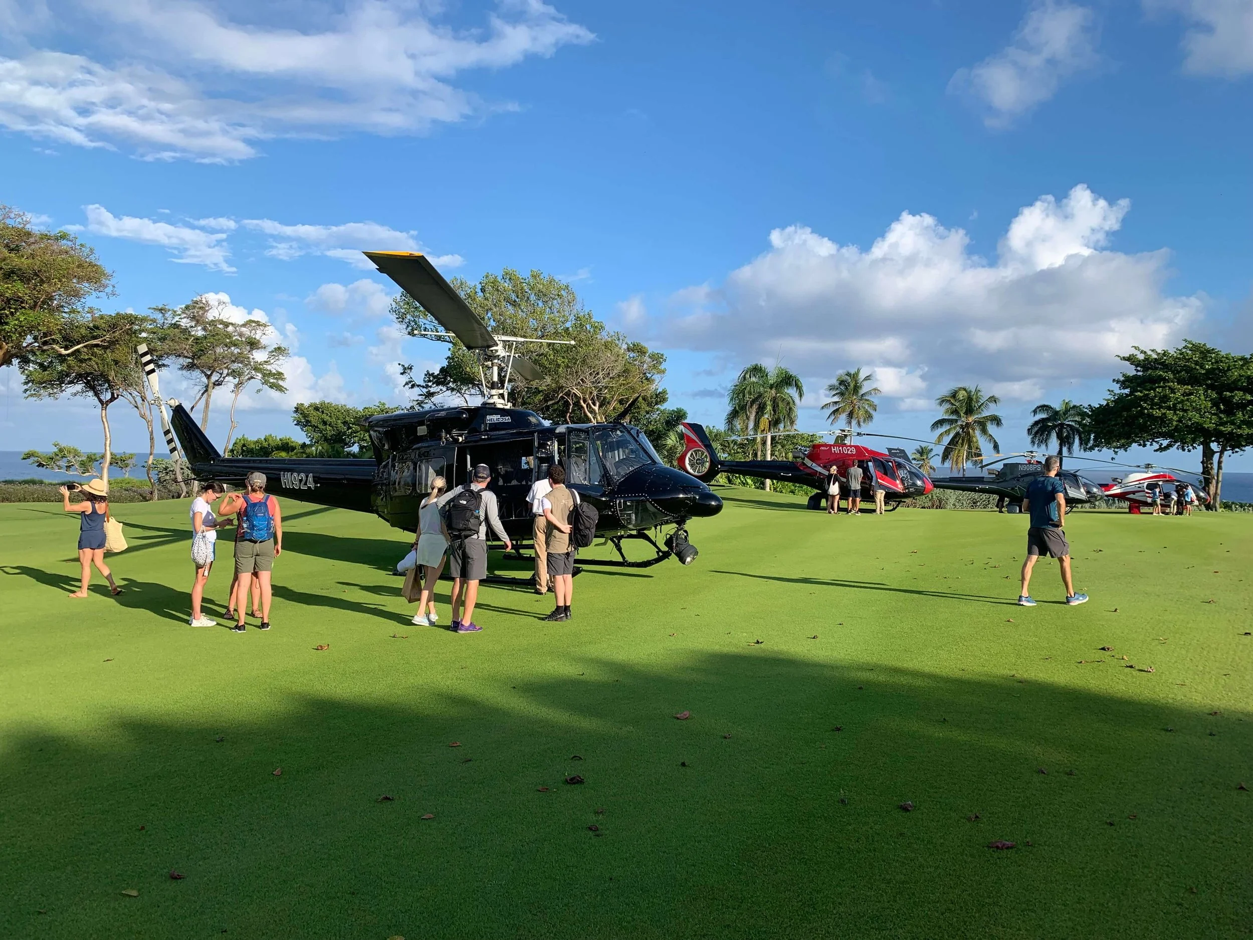 A group of people standing next to a black helicopter on a grassy field, with two other helicopters visible in the background and some trees and palm trees behind them.