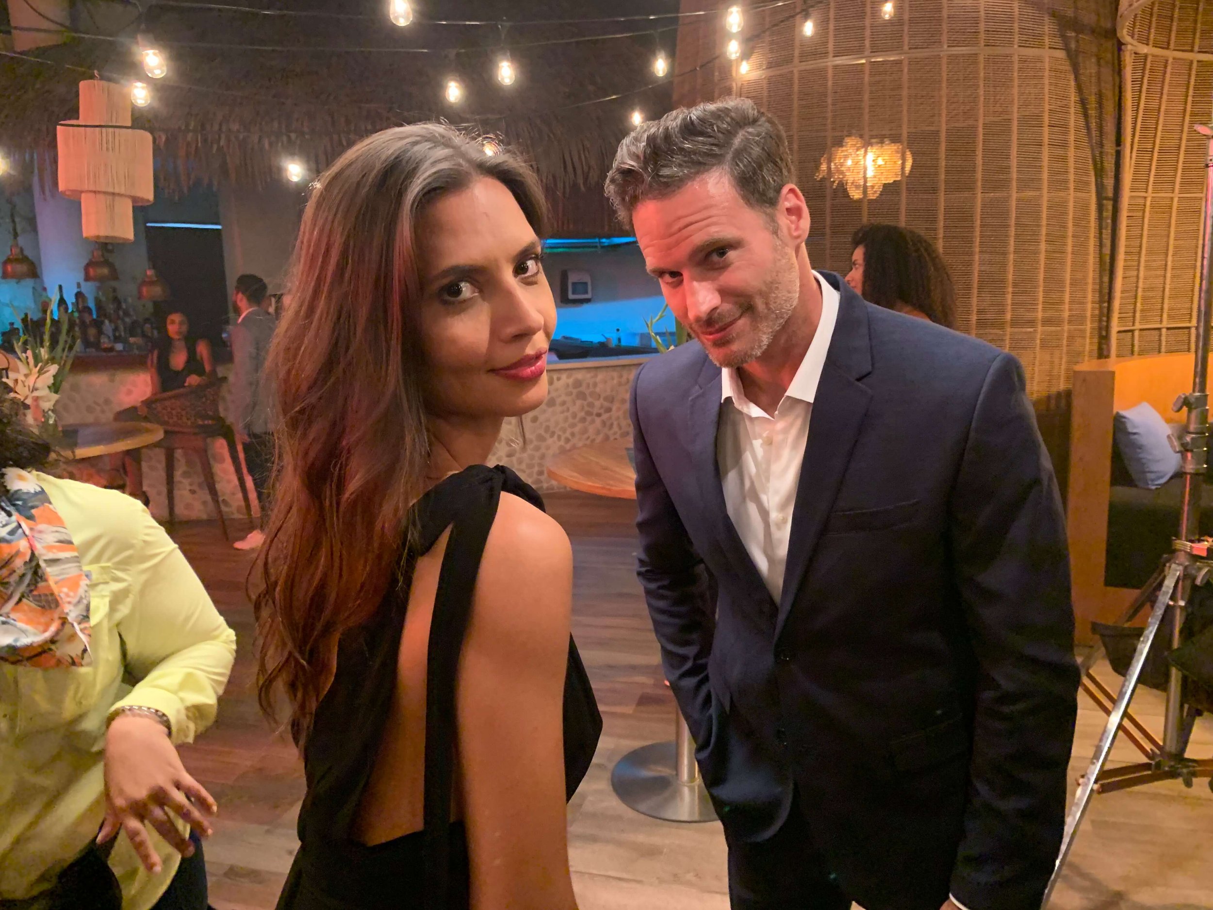 A woman with long brown hair and a man in a navy blue suit pose together at a lively indoor event with warm lighting, wooden decor, and other guests in the background.