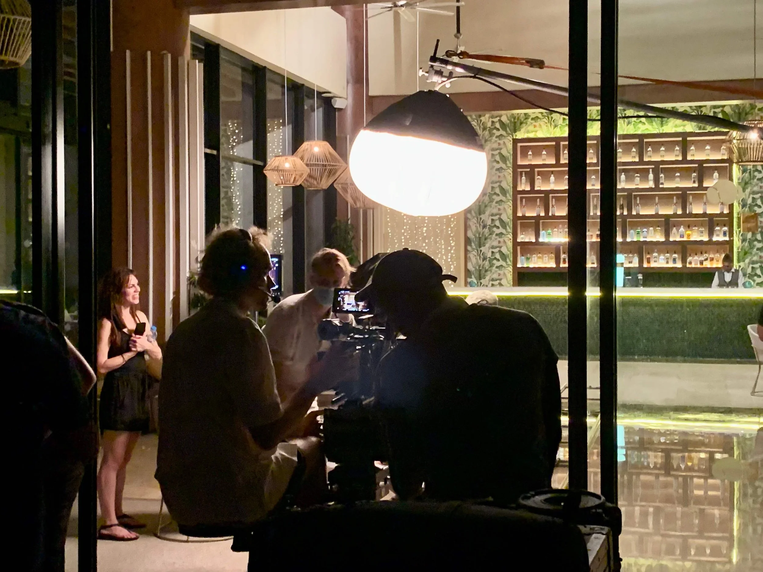 A night scene inside a bar or lounge with a camera crew filming a woman holding a drink and a cellphone, while a bartender stands behind the bar in the background. The setting features warm lighting, decorative wall with bottles, and large windows.